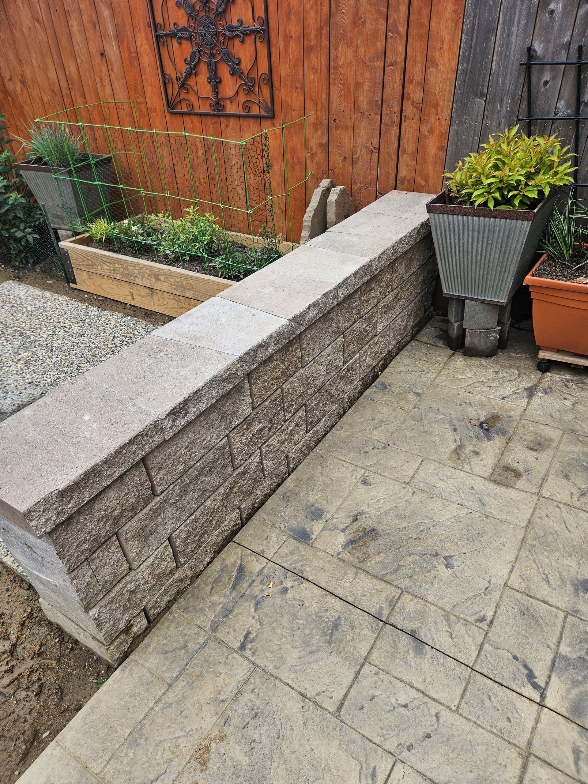 A low retaining wall made of textured stone blocks stands on a paved patio beside raised garden beds and wooden fencing.