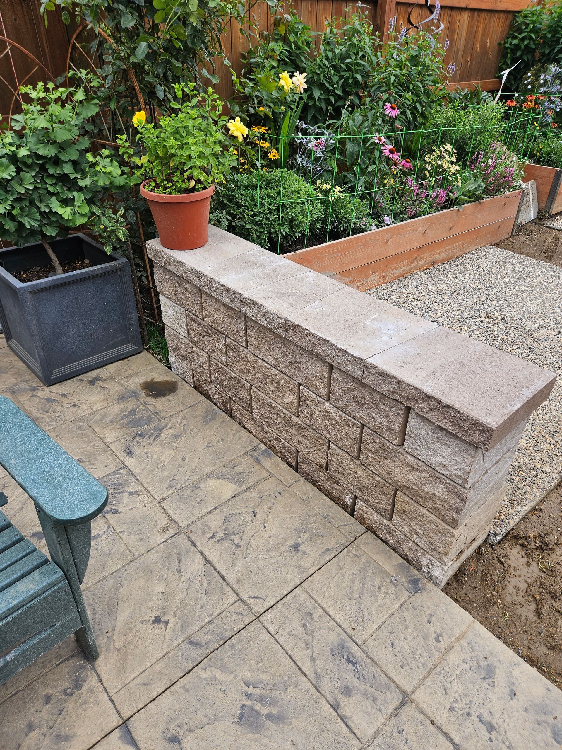A low stone retaining wall sits on a paved patio beside potted plants, a wooden garden bed, and a gravel area.