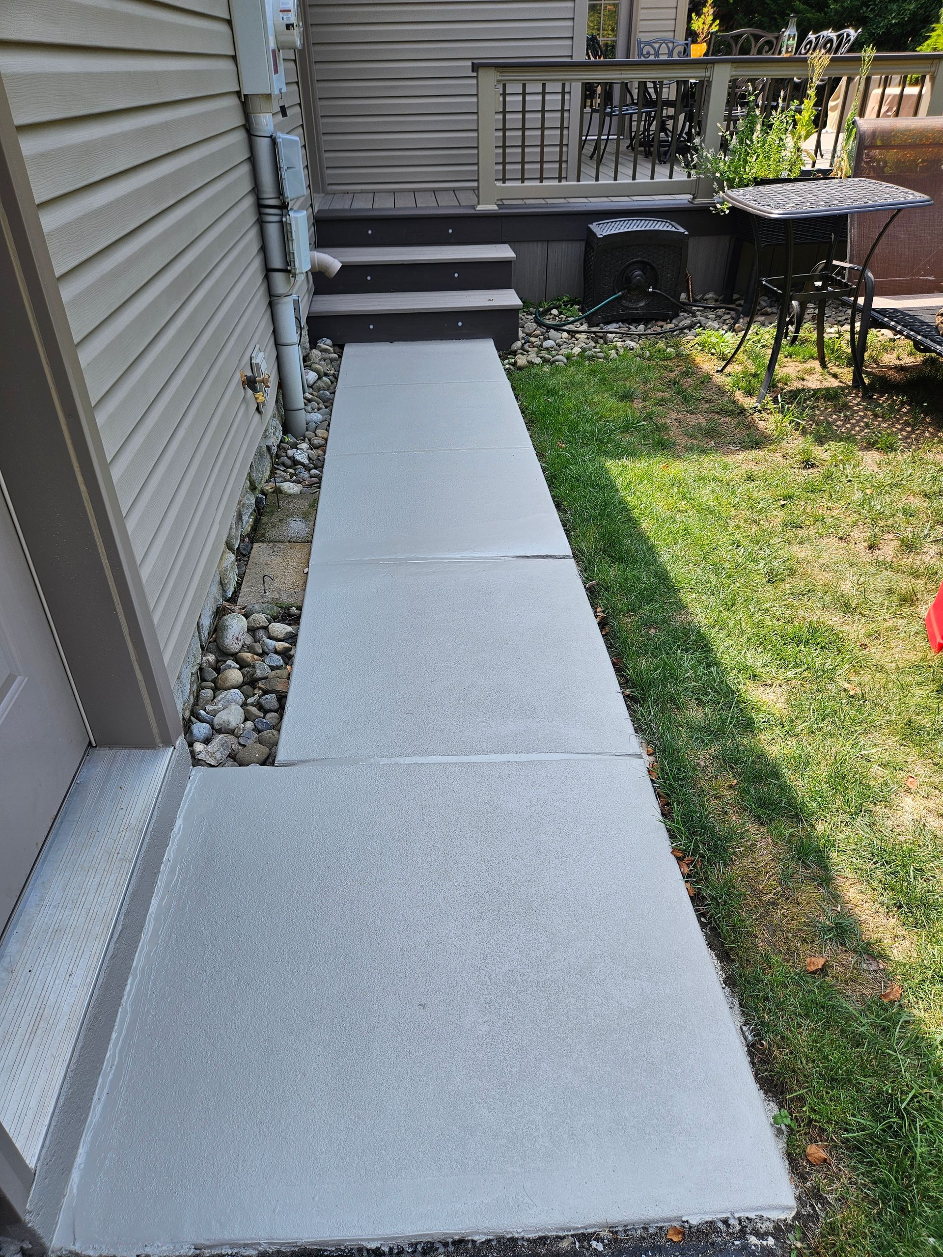 A light gray concrete walkway runs alongside a house with beige siding, leading to a small set of steps up to a deck.