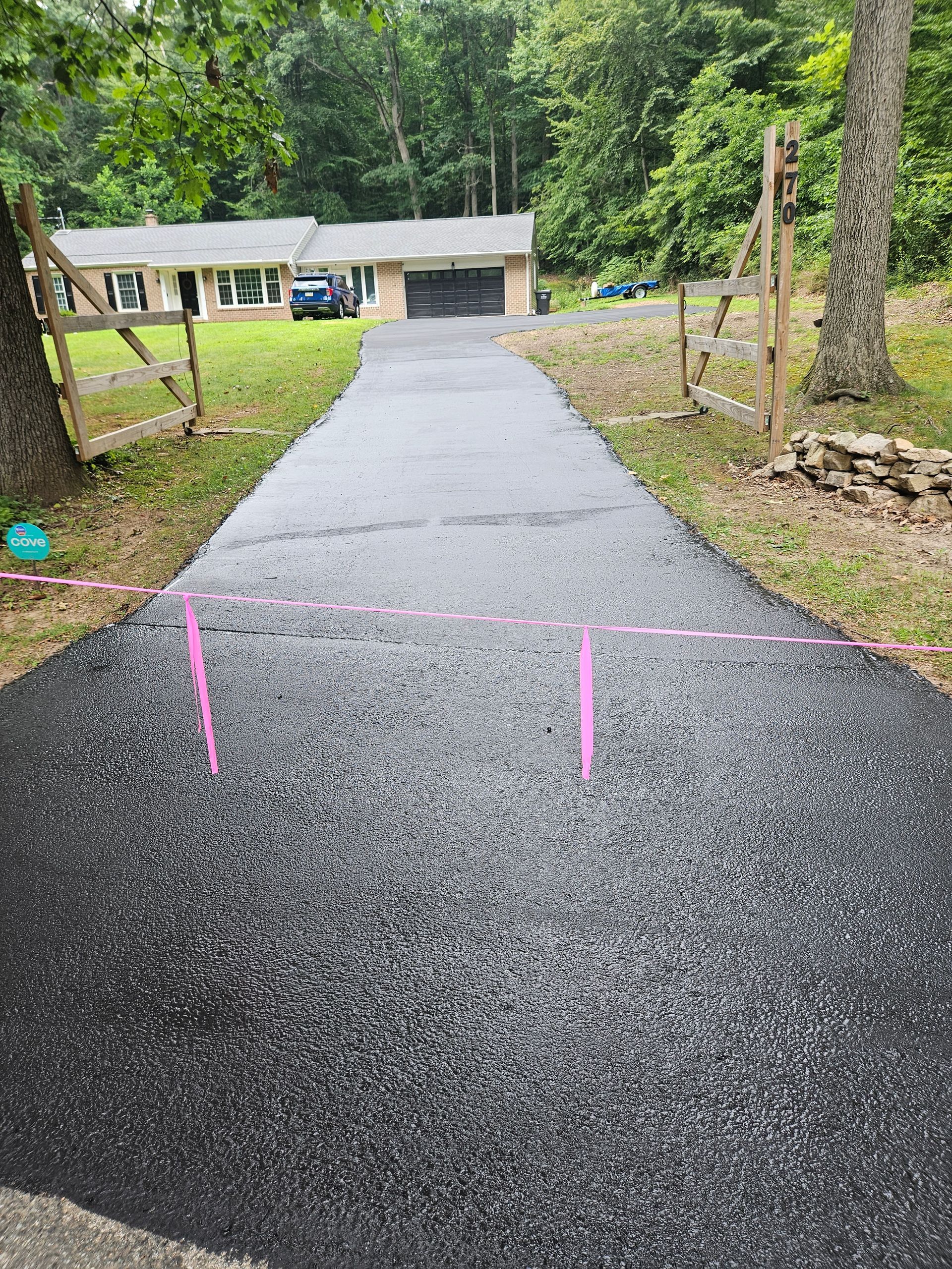 A freshly paved black asphalt driveway leads toward a one-story brick house with a garage, flanked by wooden fence posts.
