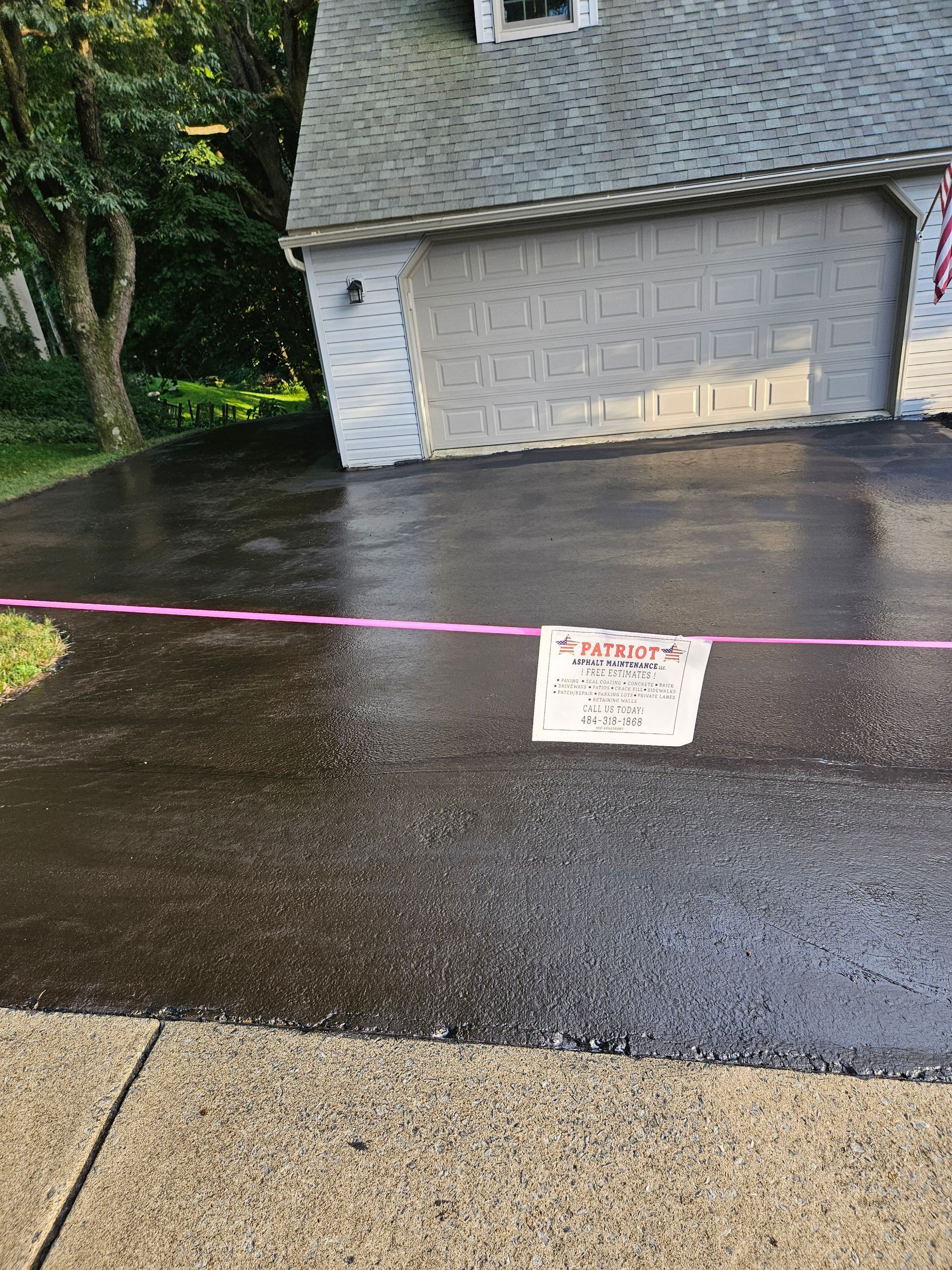 A driveway freshly coated in black asphalt, cordoned off with pink caution tape and a handwritten sign.