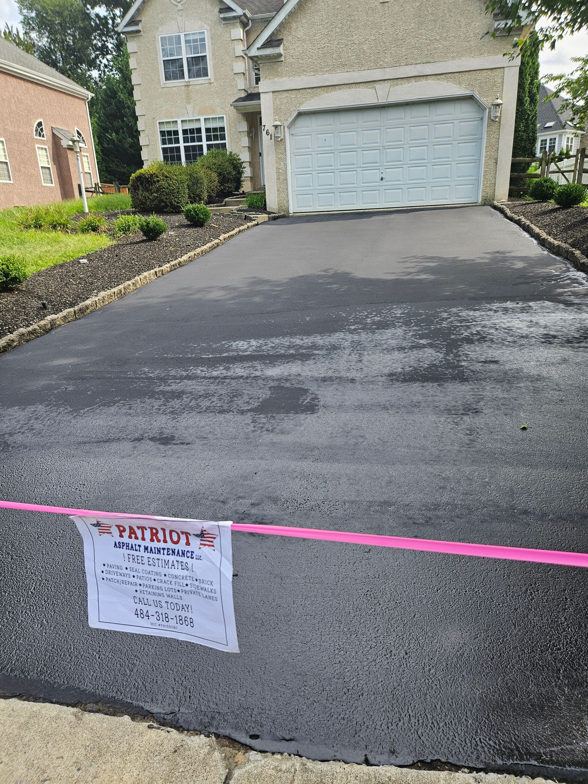 A newly paved asphalt driveway leads to a suburban home, blocked by a pink caution tape with a sign attached.