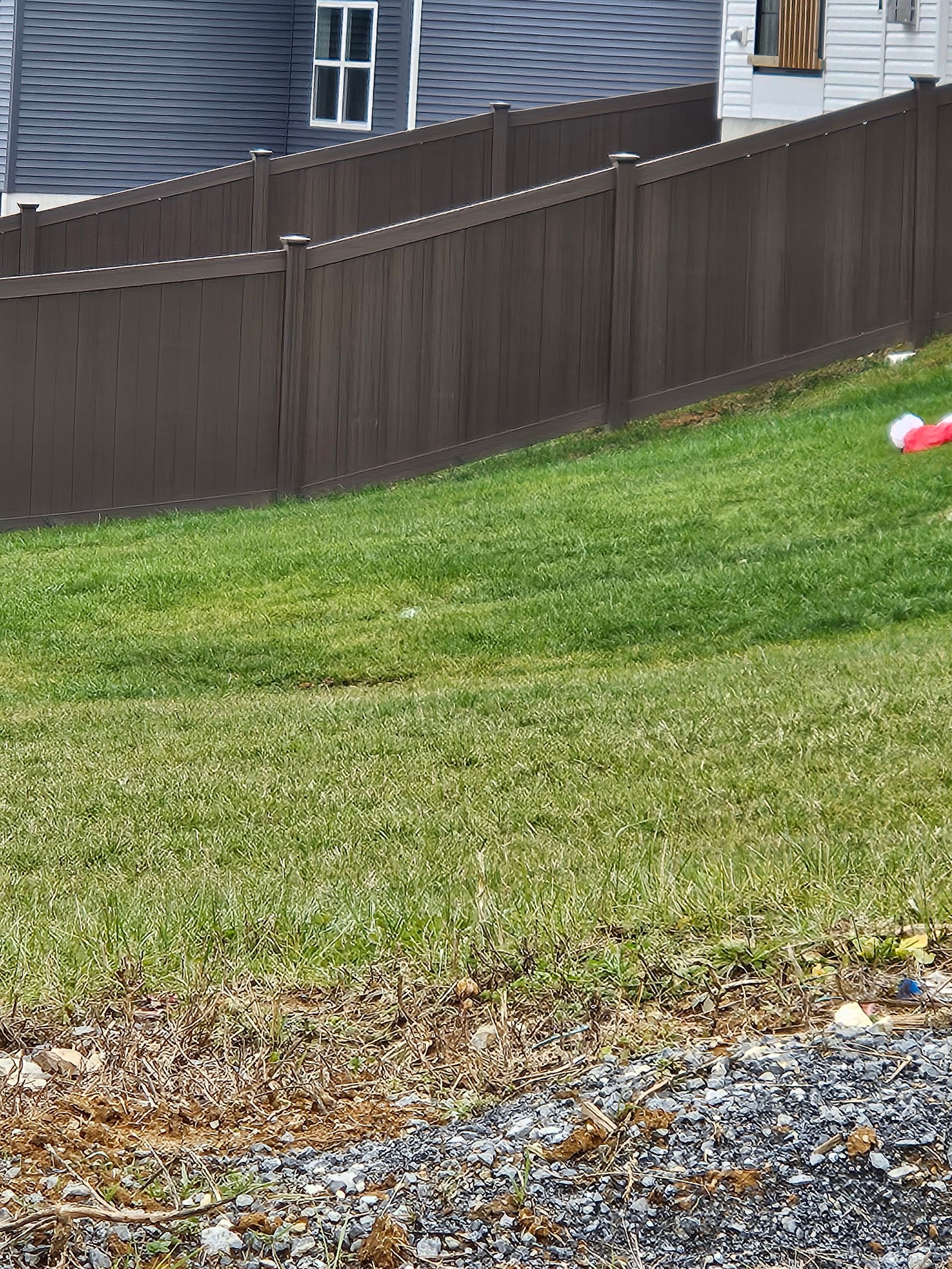 A brown privacy fence runs diagonally across a green, grassy sloped lawn, with a partial house visible in the background.