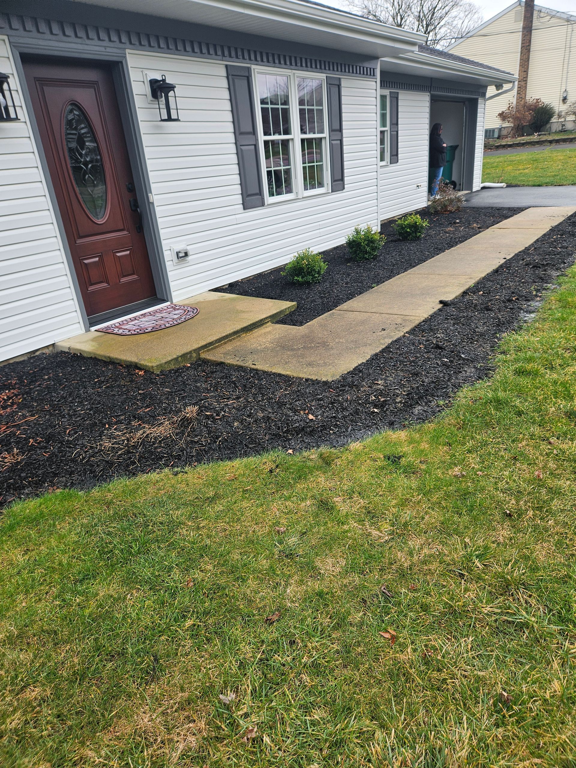 A white house with gray shutters, a brown front door, and a concrete path surrounded by black mulch.