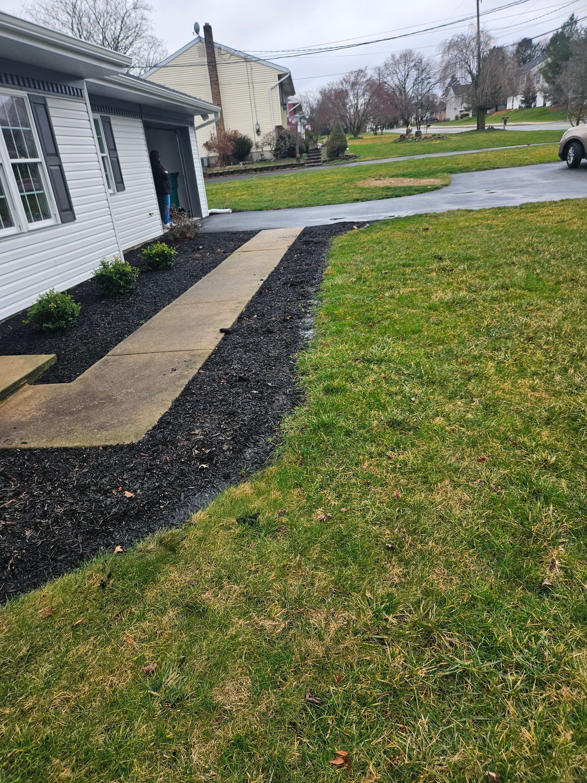 A house exterior with a concrete sidewalk bordered by fresh black mulch and a green grass lawn under a cloudy sky.