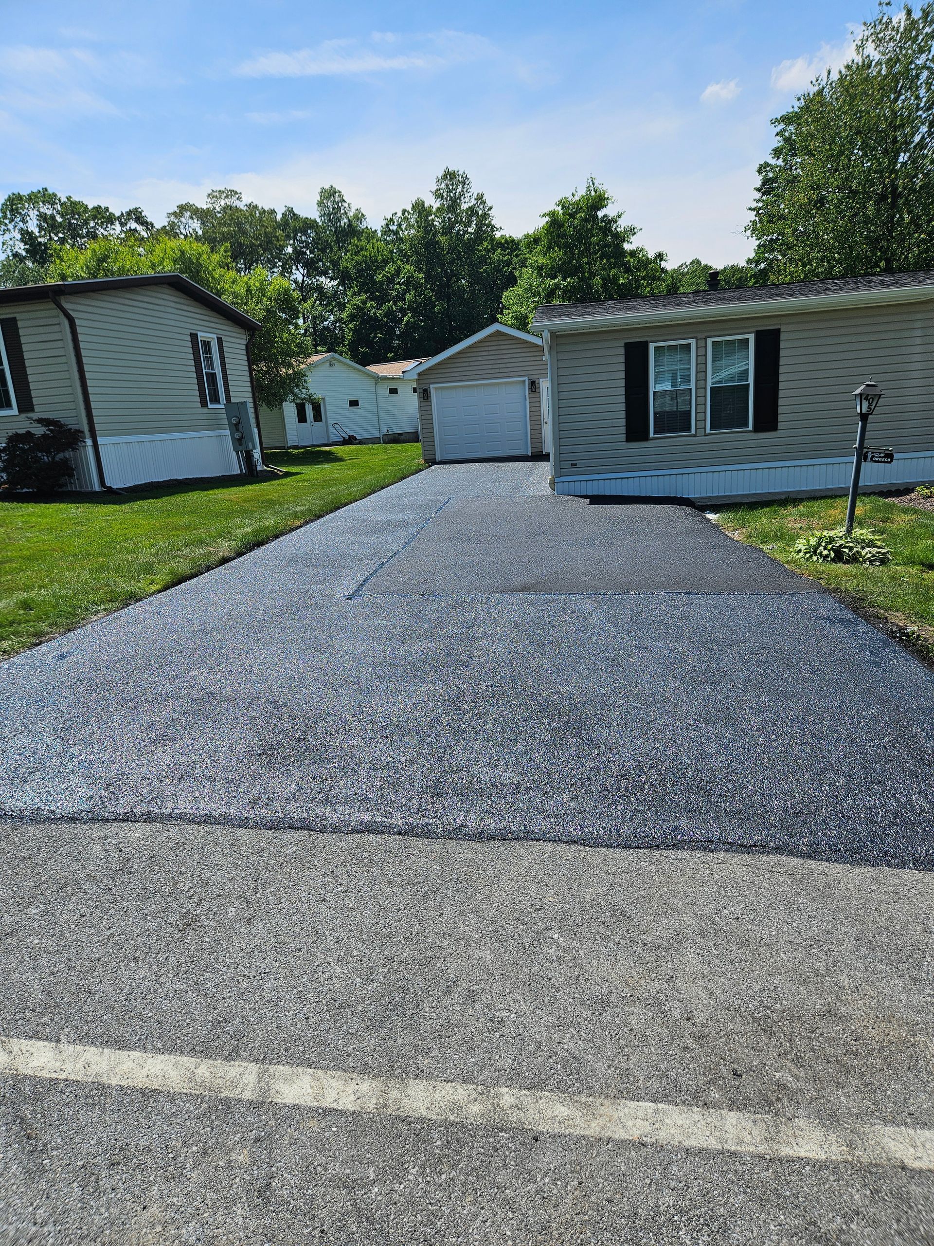 A newly laid, dark gravel driveway extends between two manufactured homes toward a detached garage under a sunny sky.