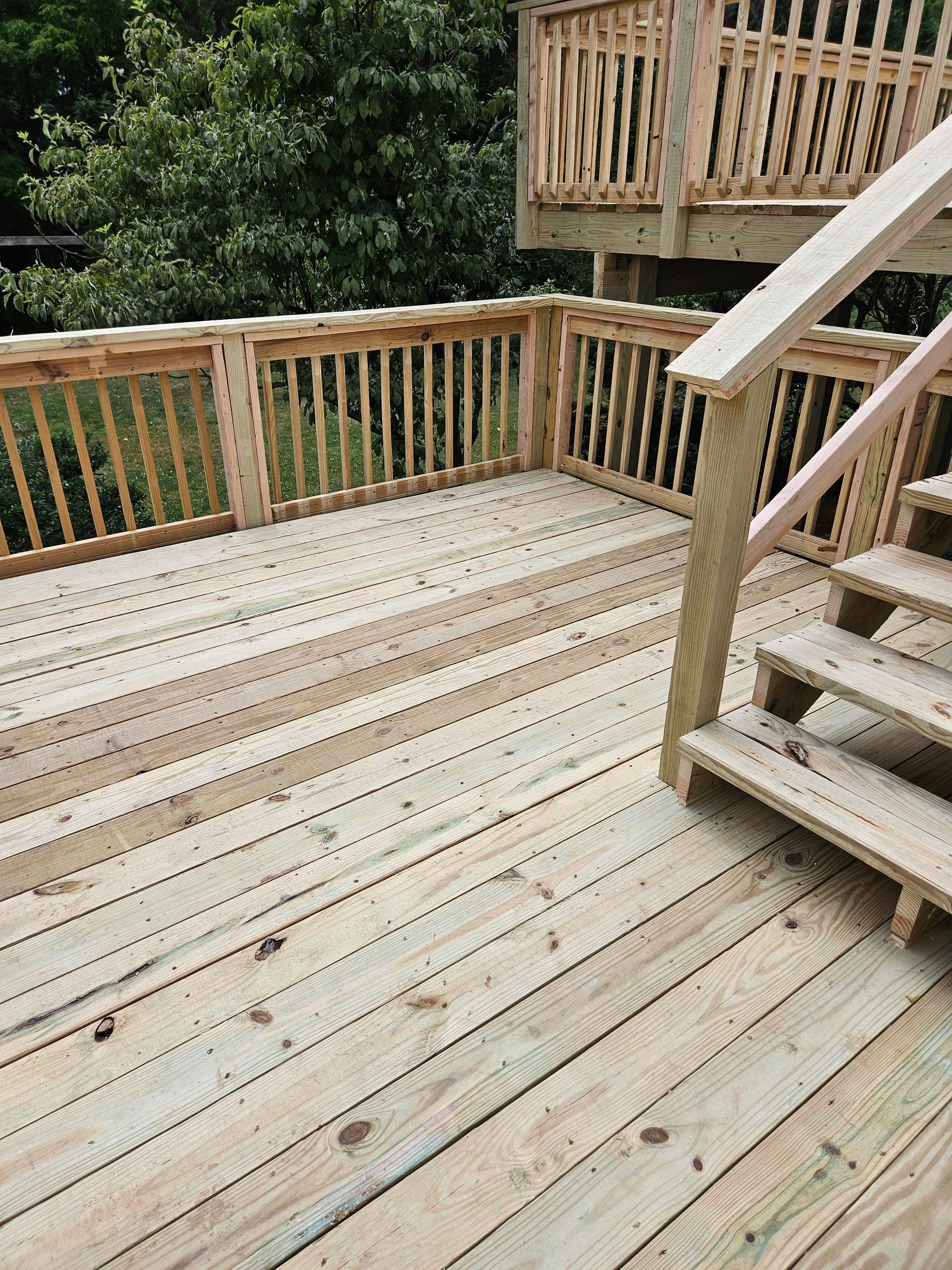 A light-colored wooden deck with a railing and staircase leading to a higher level, set outdoors against a tree.