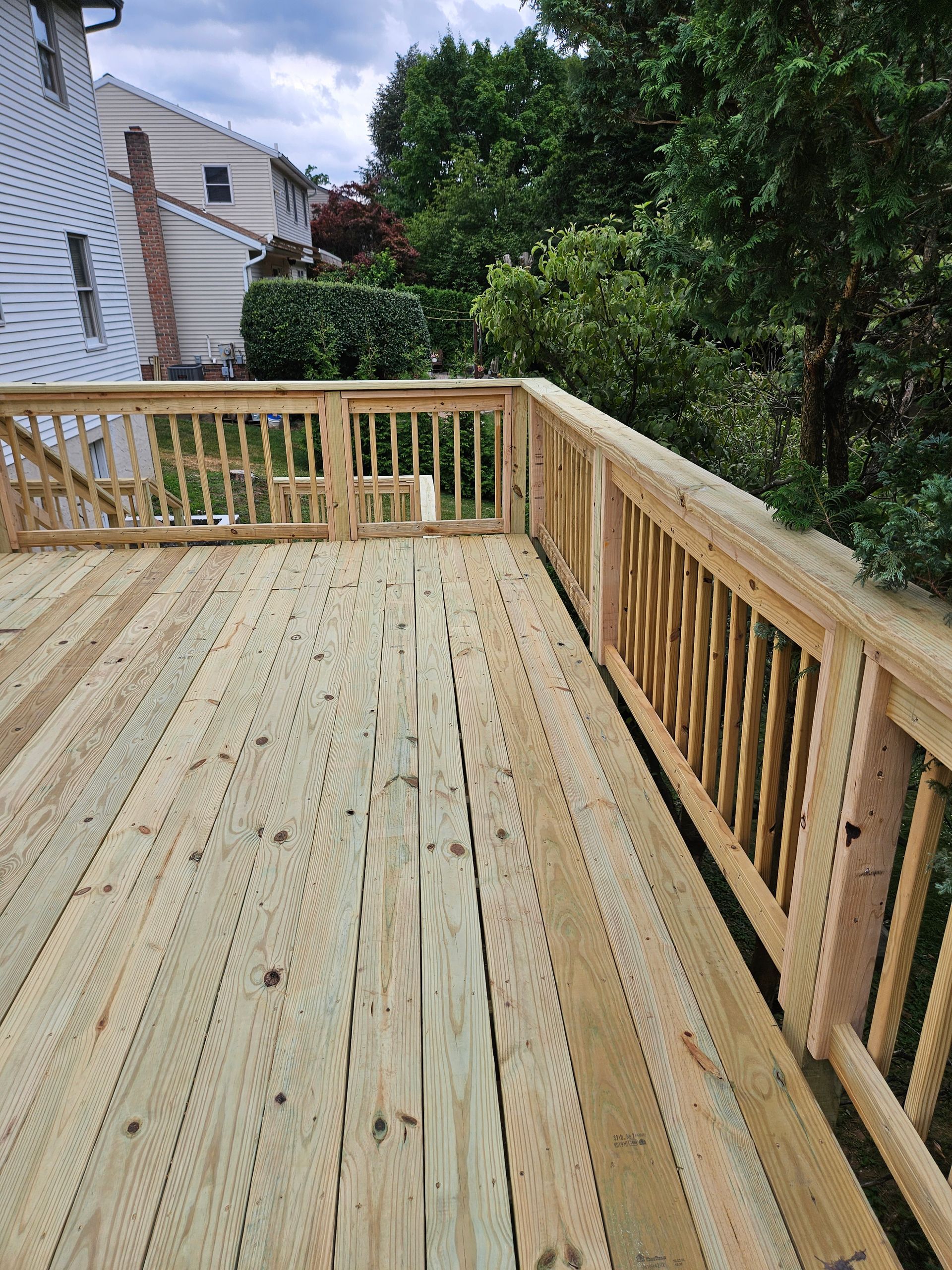 A newly built wooden deck with railings, viewed from above, overlooking a backyard with green trees and a nearby house.
