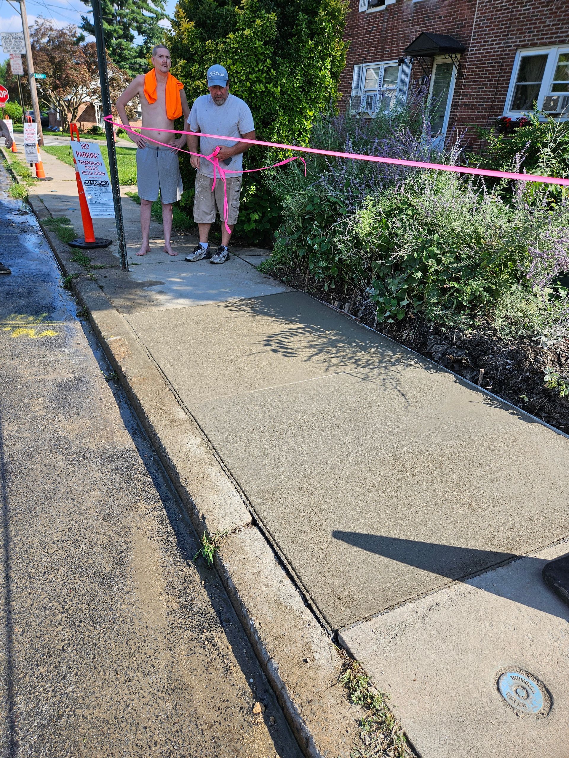Two people stand behind a pink caution tape cordoning off a freshly poured concrete sidewalk section near a brick building.
