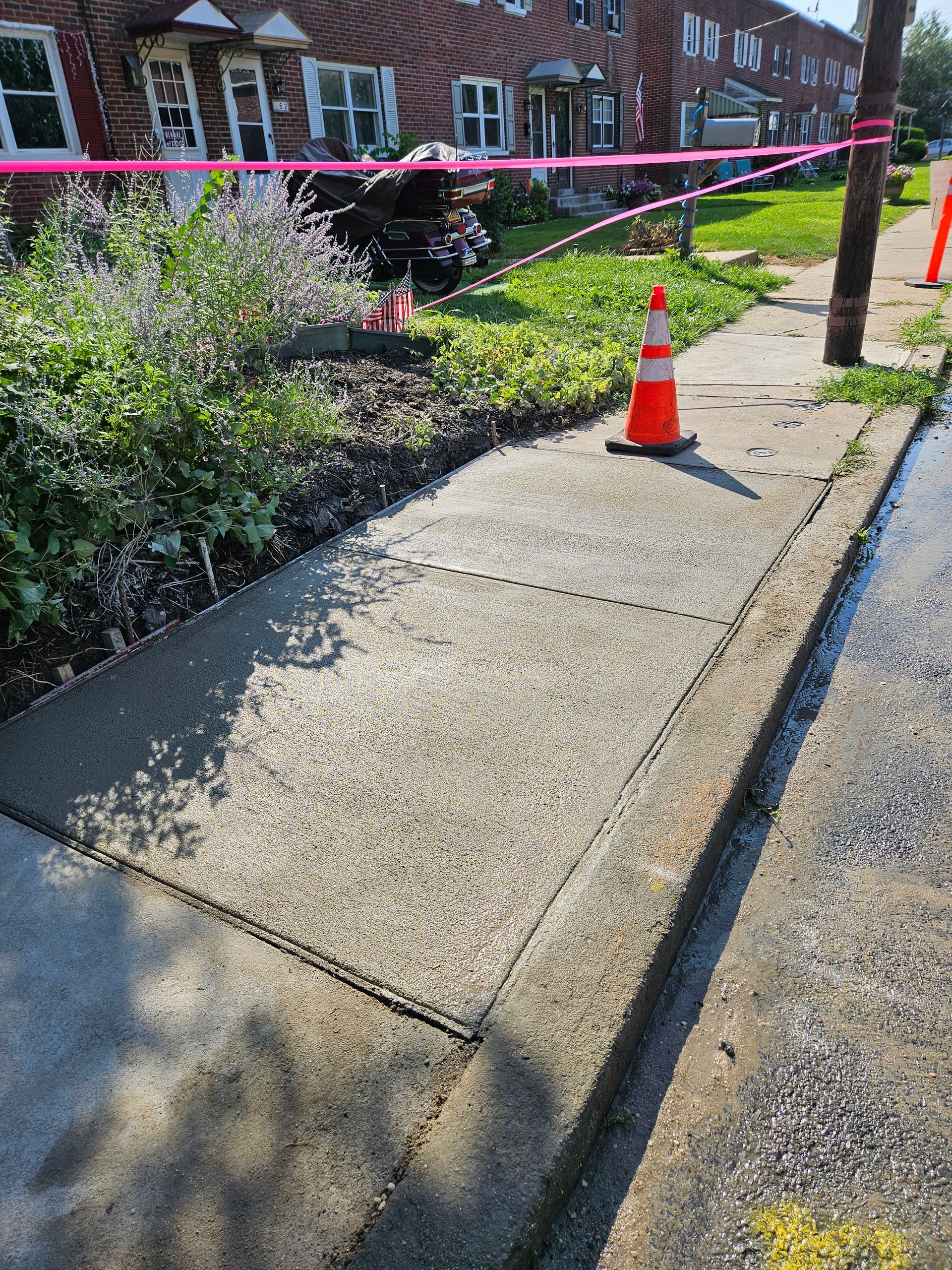A sidewalk in front of a brick building, blocked by a bright orange traffic cone and pink caution tape.