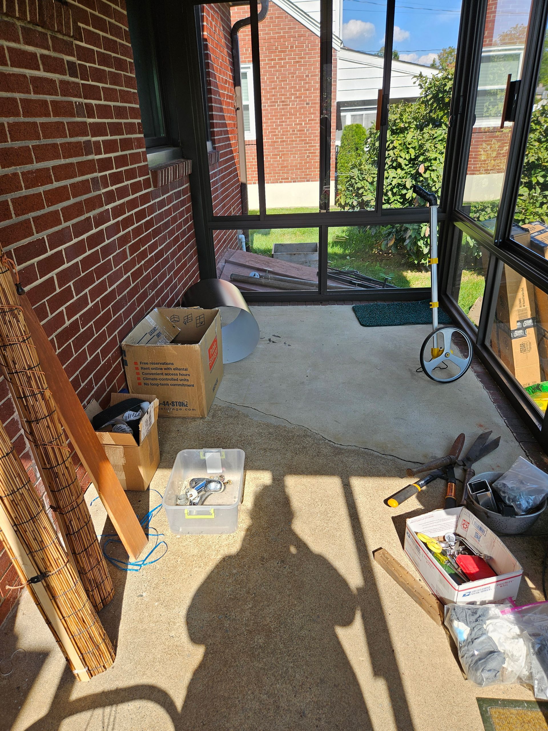 A sunroom with a brick wall, concrete floor, scattered cardboard boxes, tools, and a measuring wheel by the screen door.