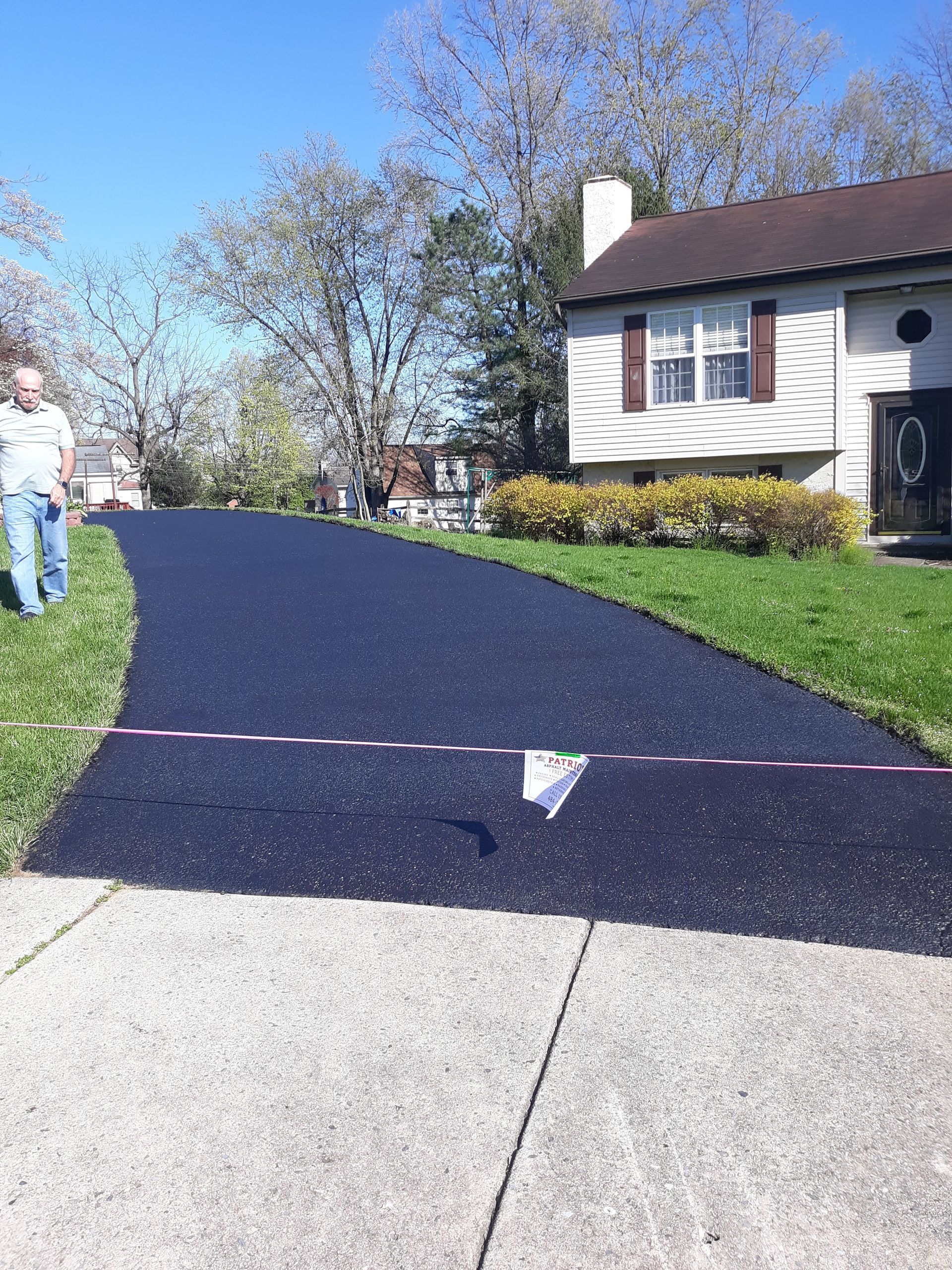A person stands near a freshly paved black asphalt driveway that curves up toward a house on a sunny day.