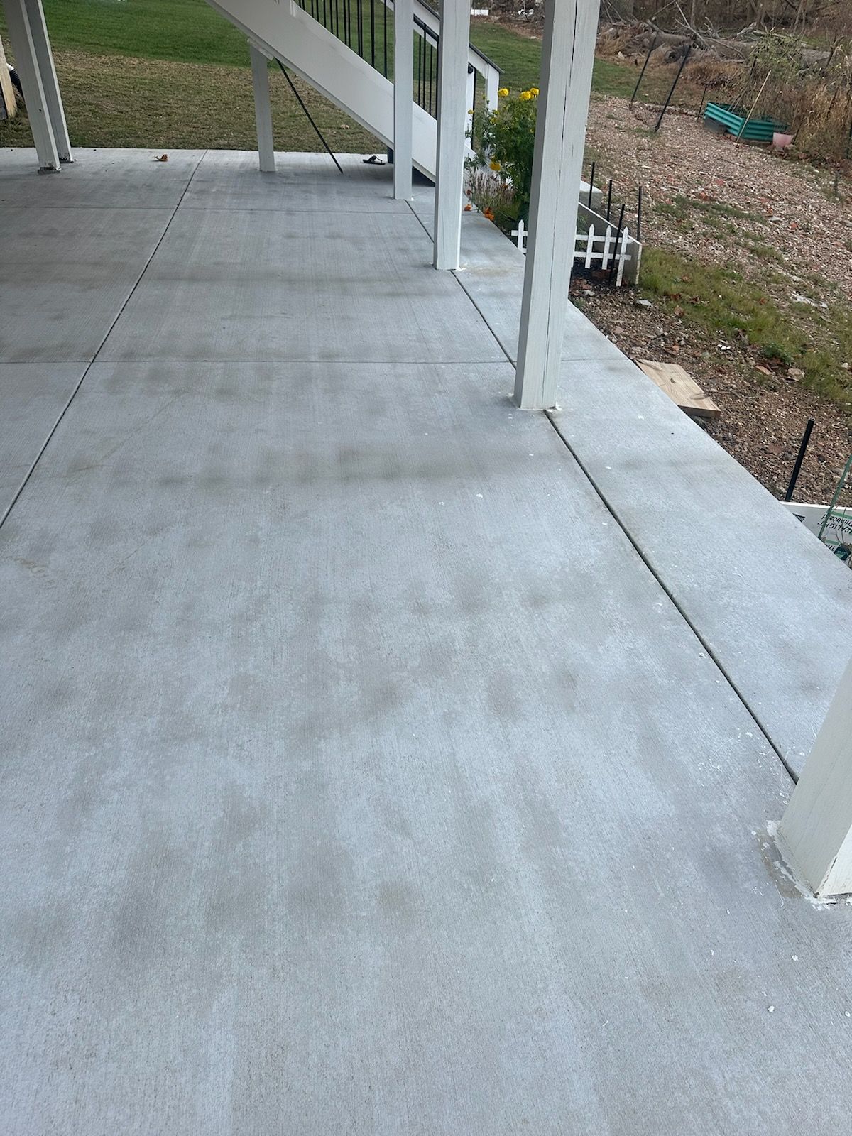 A gray concrete patio surface with support posts for an elevated deck above, bordered by grass and a garden area.