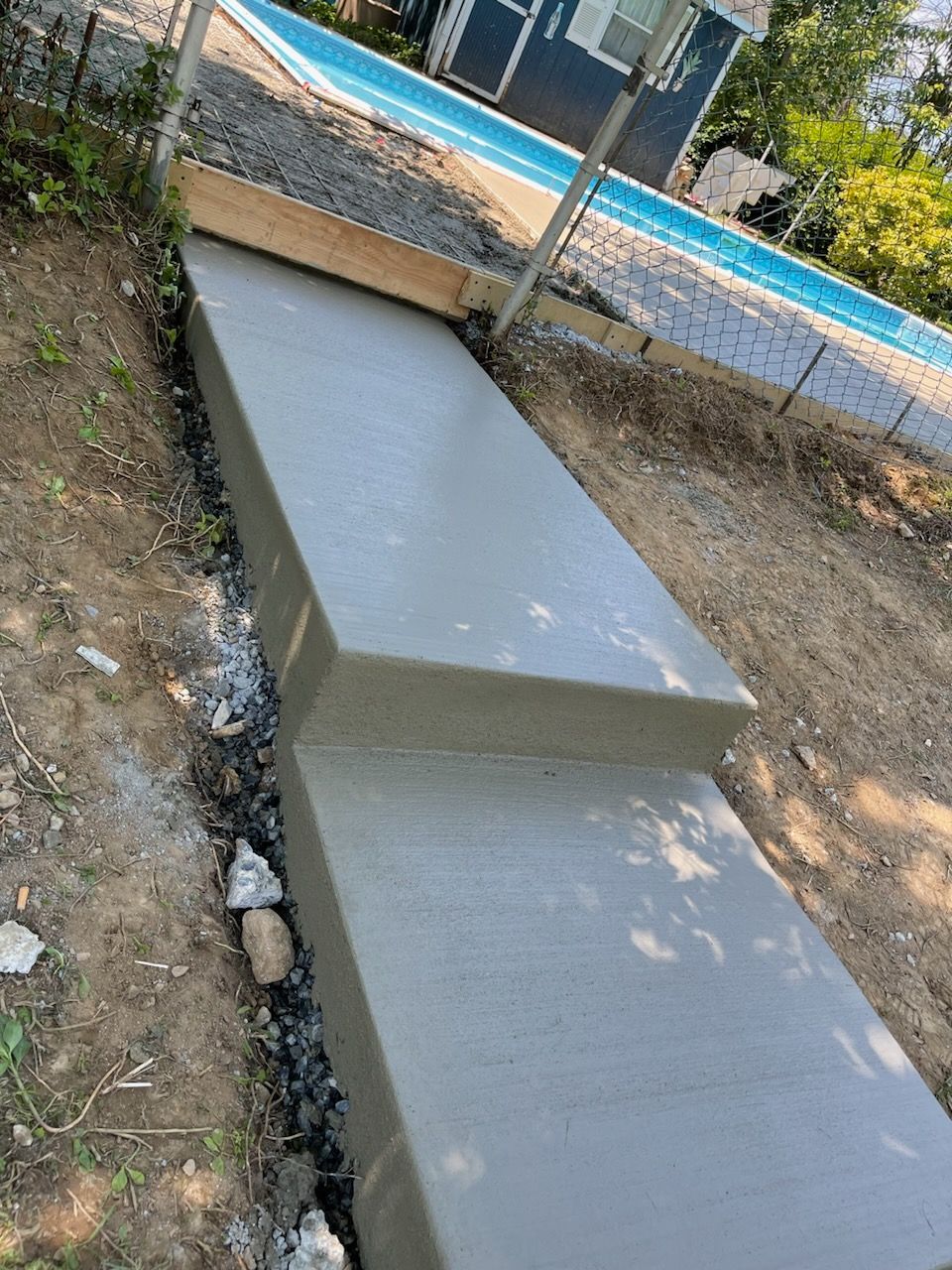 Two freshly poured concrete steps leading up to a wooden-framed area next to a blue swimming pool.