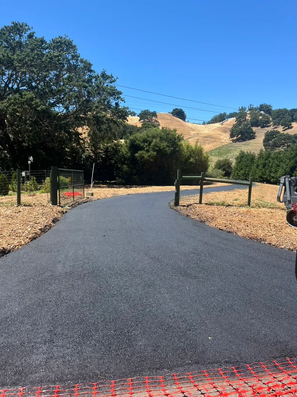 New black asphalt path curving through a dry hillside with trees and posts on both sides