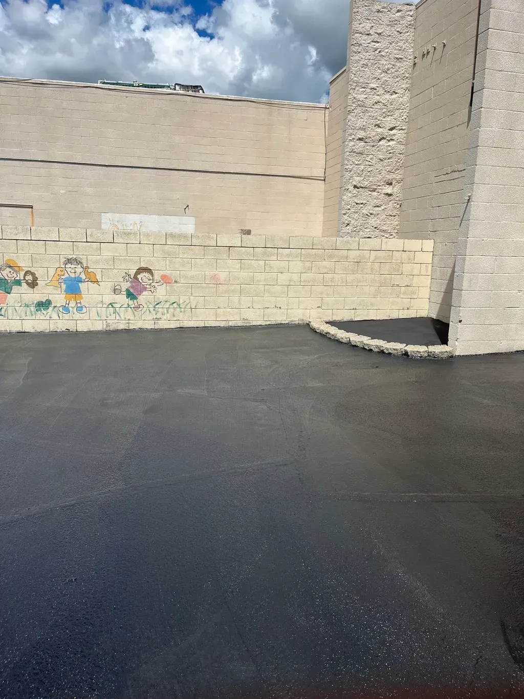 Empty paved courtyard with tan block walls, a mural, and a partly cloudy sky.
