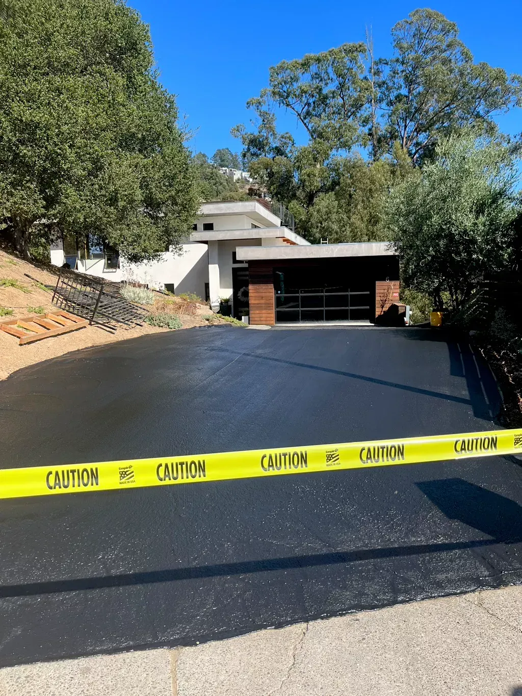 Caution tape blocks a steep driveway leading to a house garage, with wet black pavement and trees overhead.