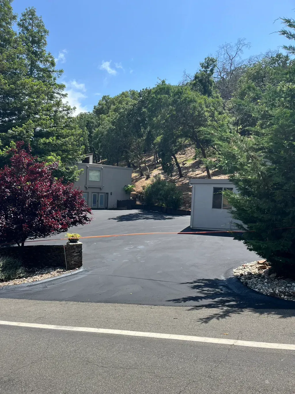 Driveway entrance to a home with trees, asphalt, and a small building under a sunny blue sky