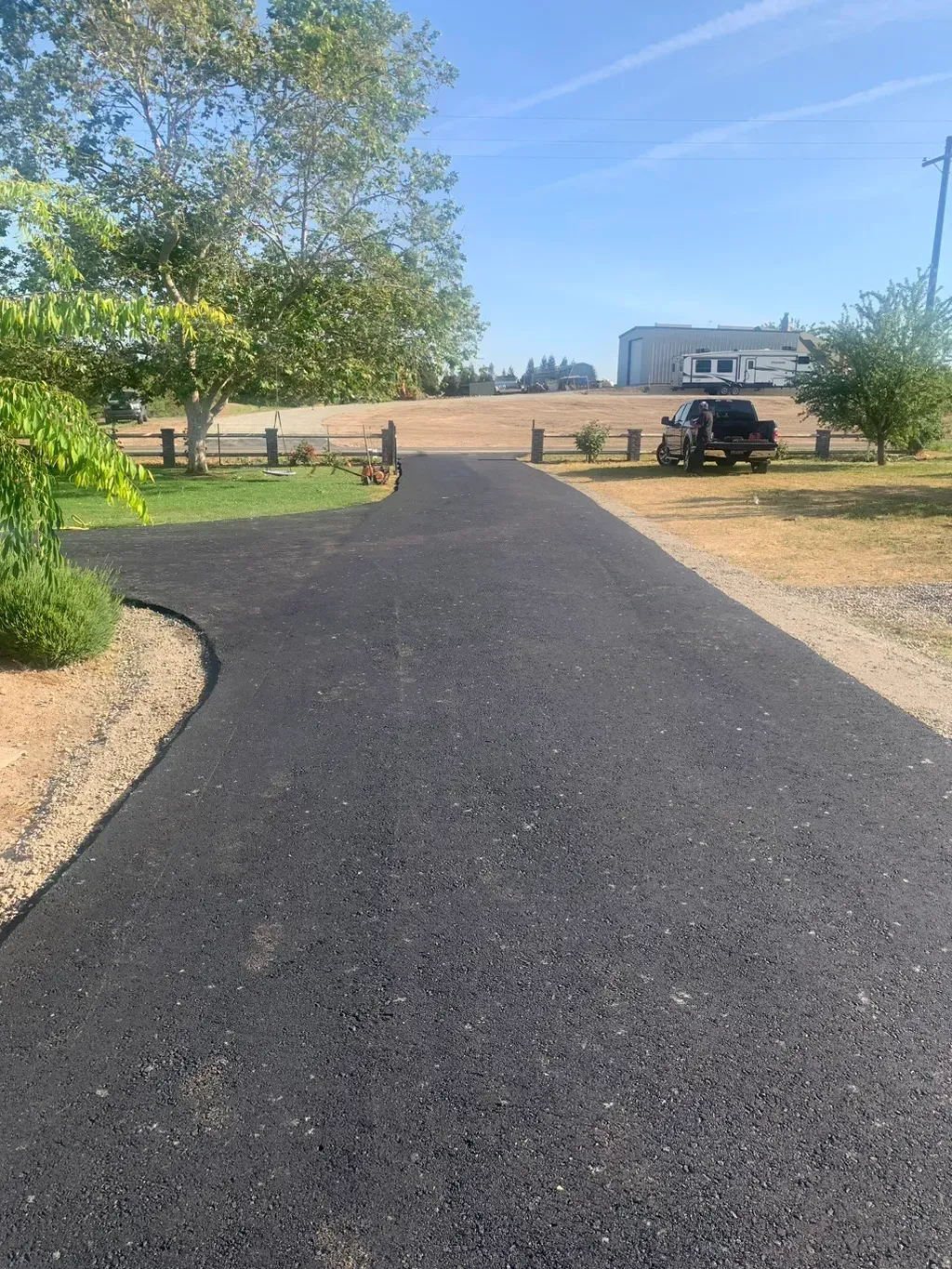 Paved driveway curving through a dry rural yard with trees, fence, and a parked vehicle near a house.