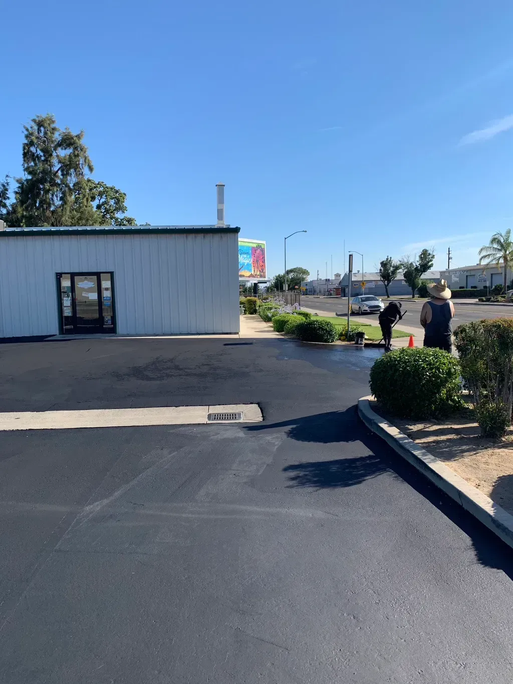 Parking lot beside a small white building, with two people walking near the curb on a sunny day.