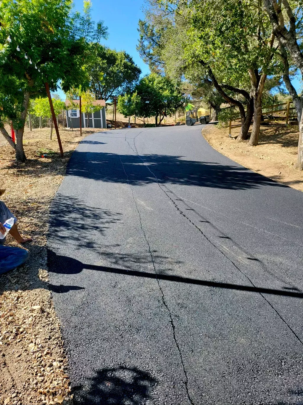 Newly paved road through a dry, tree-lined rural area with shadows across the asphalt.