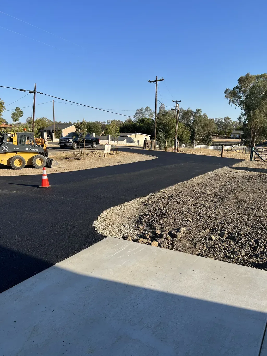 Freshly paved road under construction beside a concrete sidewalk and gravel shoulder, with cones and equipment under clear sky