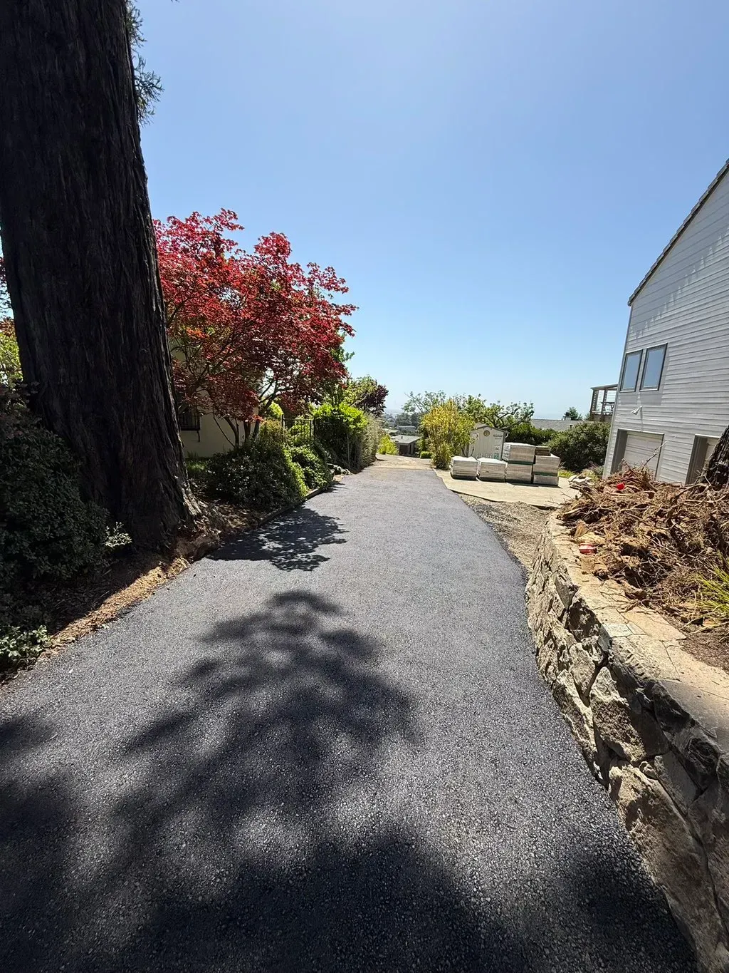 Curved gravel garden path beside a house, with shrubs, a tree, and ocean view under a bright blue sky