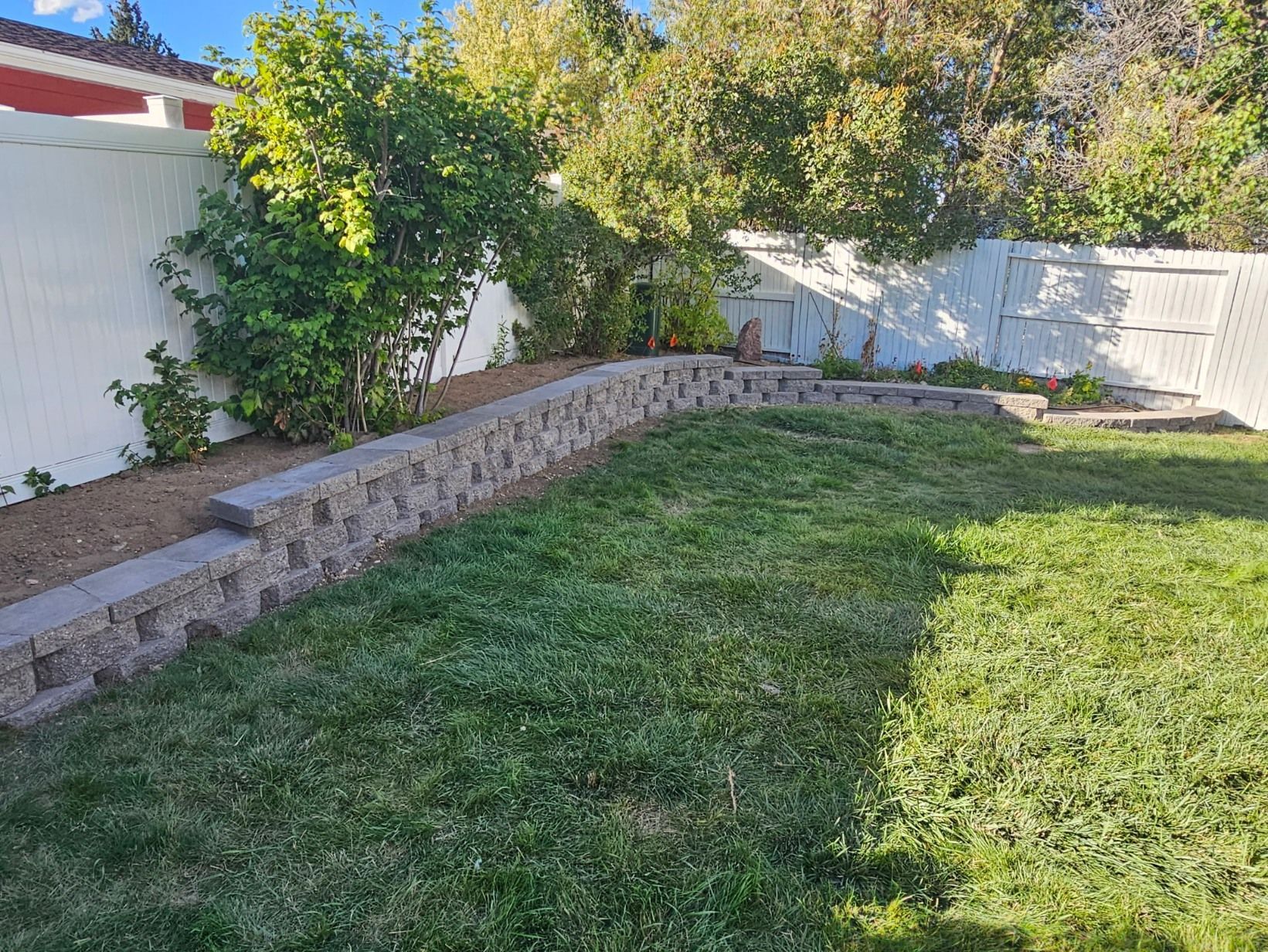 A stone retaining wall curves through a grassy backyard in front of a white fence and lush green bushes.