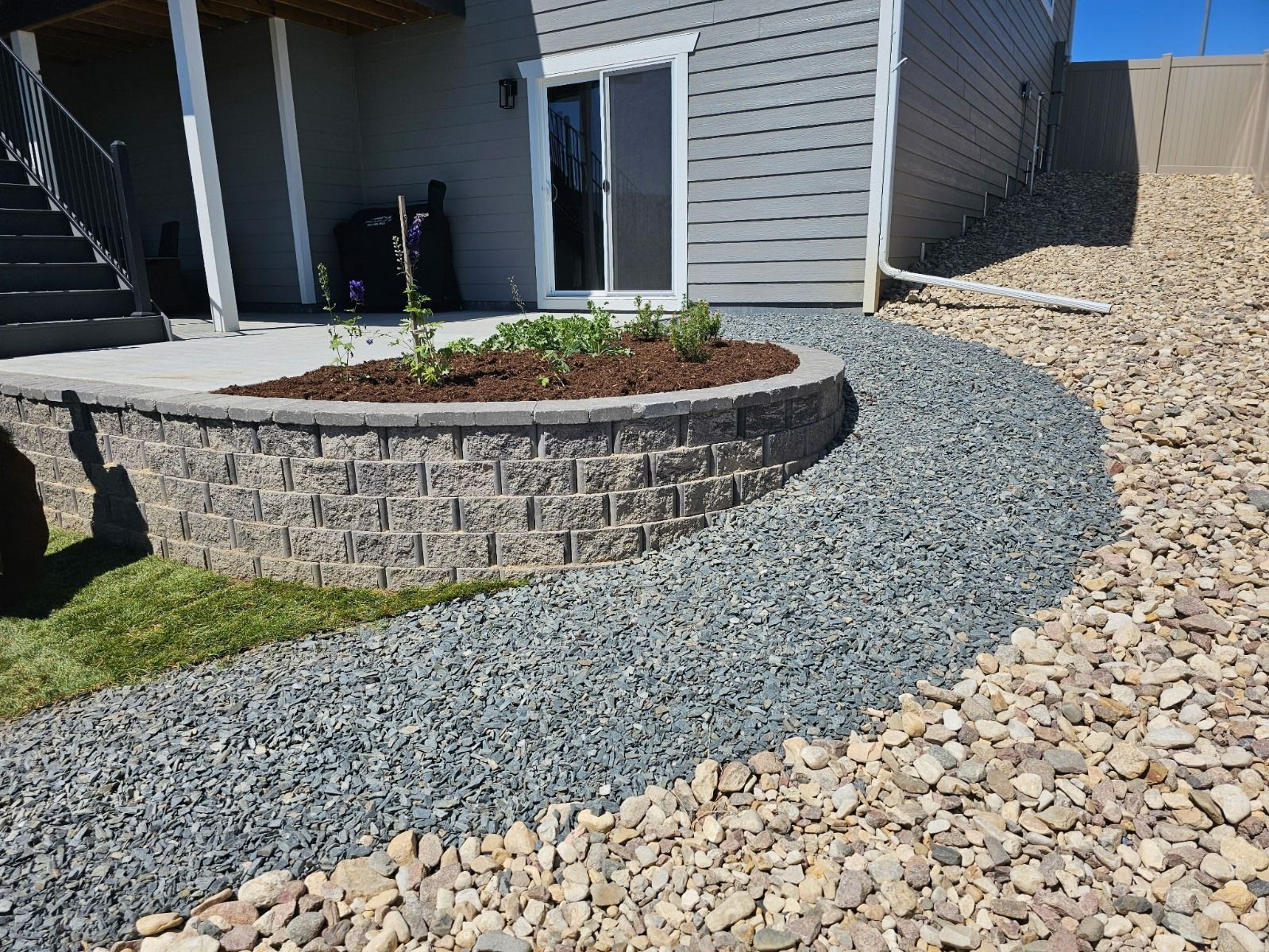 A curved stone retaining wall holds a small flower bed next to a gray house with a patio and gravel landscaping.