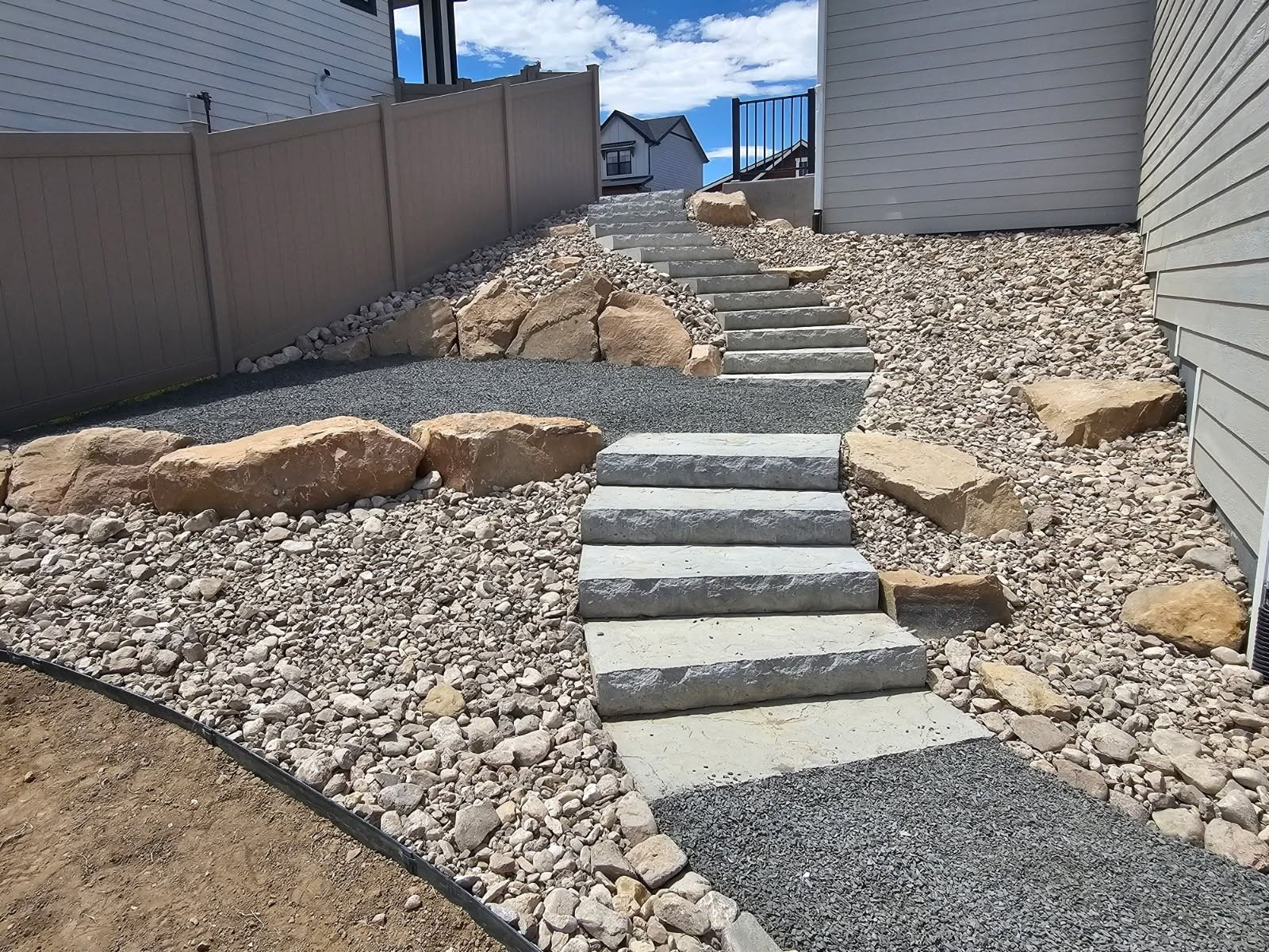 A stone staircase leads up a rocky, landscaped incline between a tan fence and the light gray siding of a house.