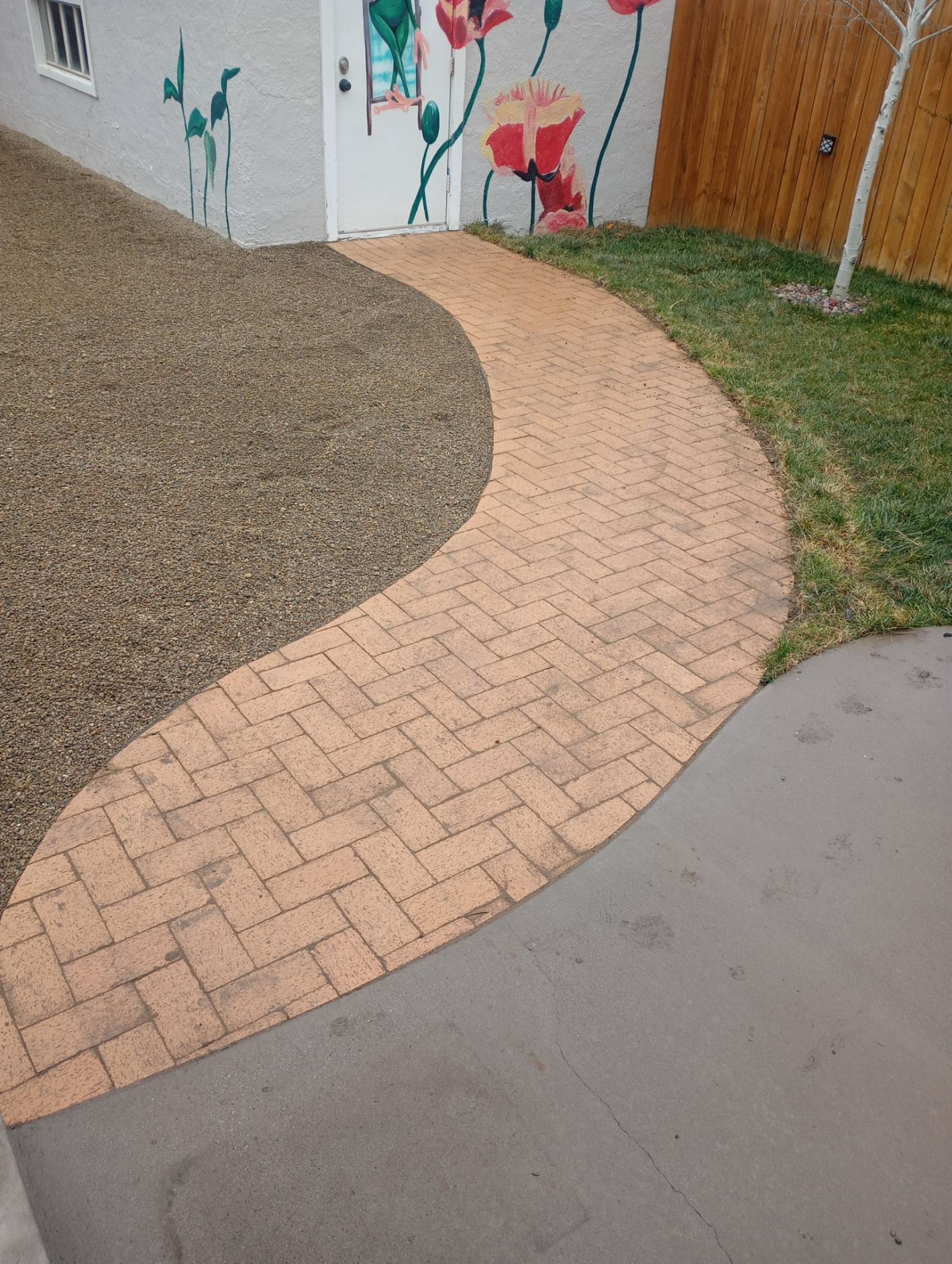 A curved brick walkway leads to a white door featuring a floral mural, flanked by gravel and a patch of grass.