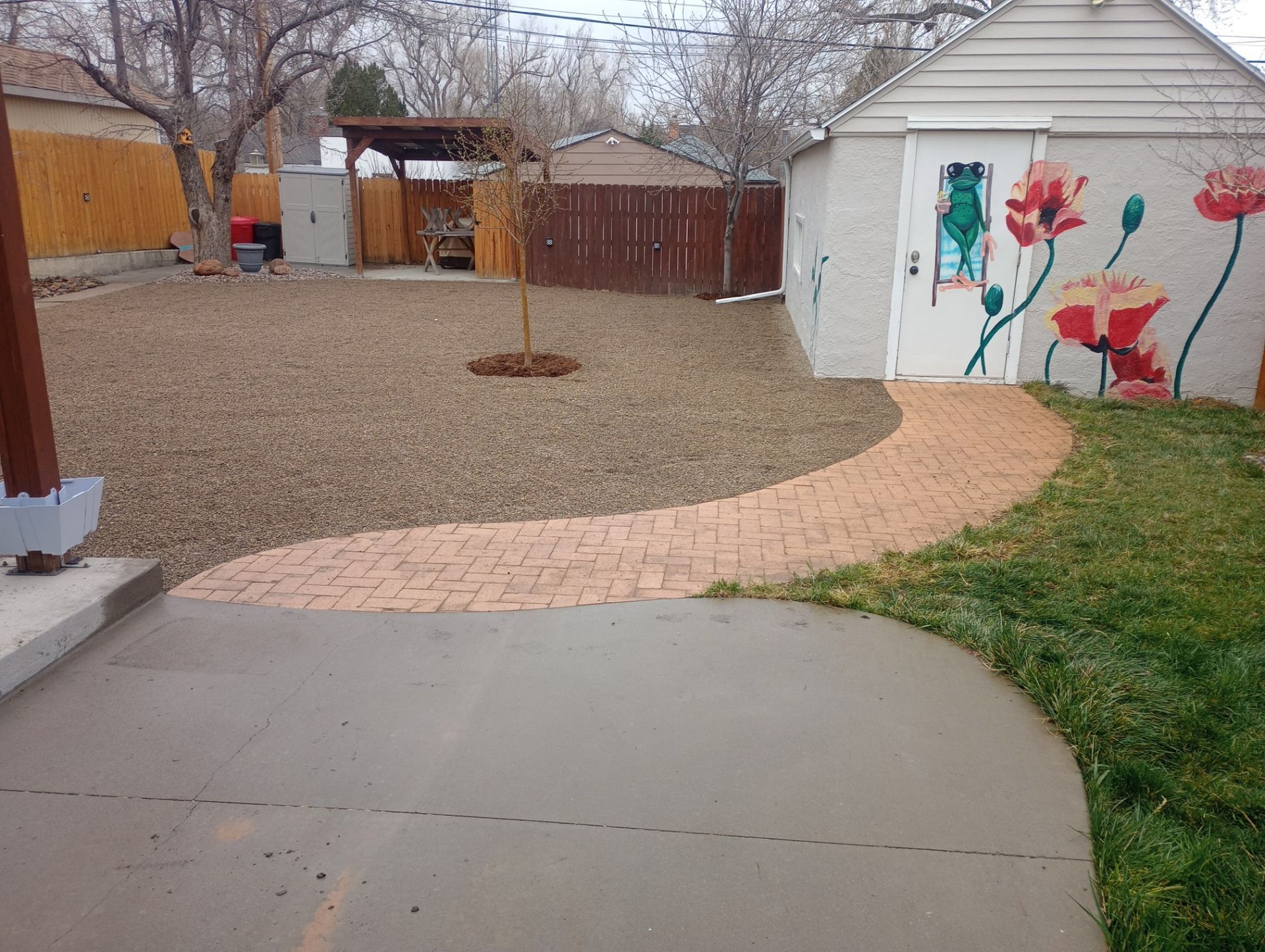 A backyard view with a concrete patio, a winding brick path, gravel yard, and a shed painted with floral murals.