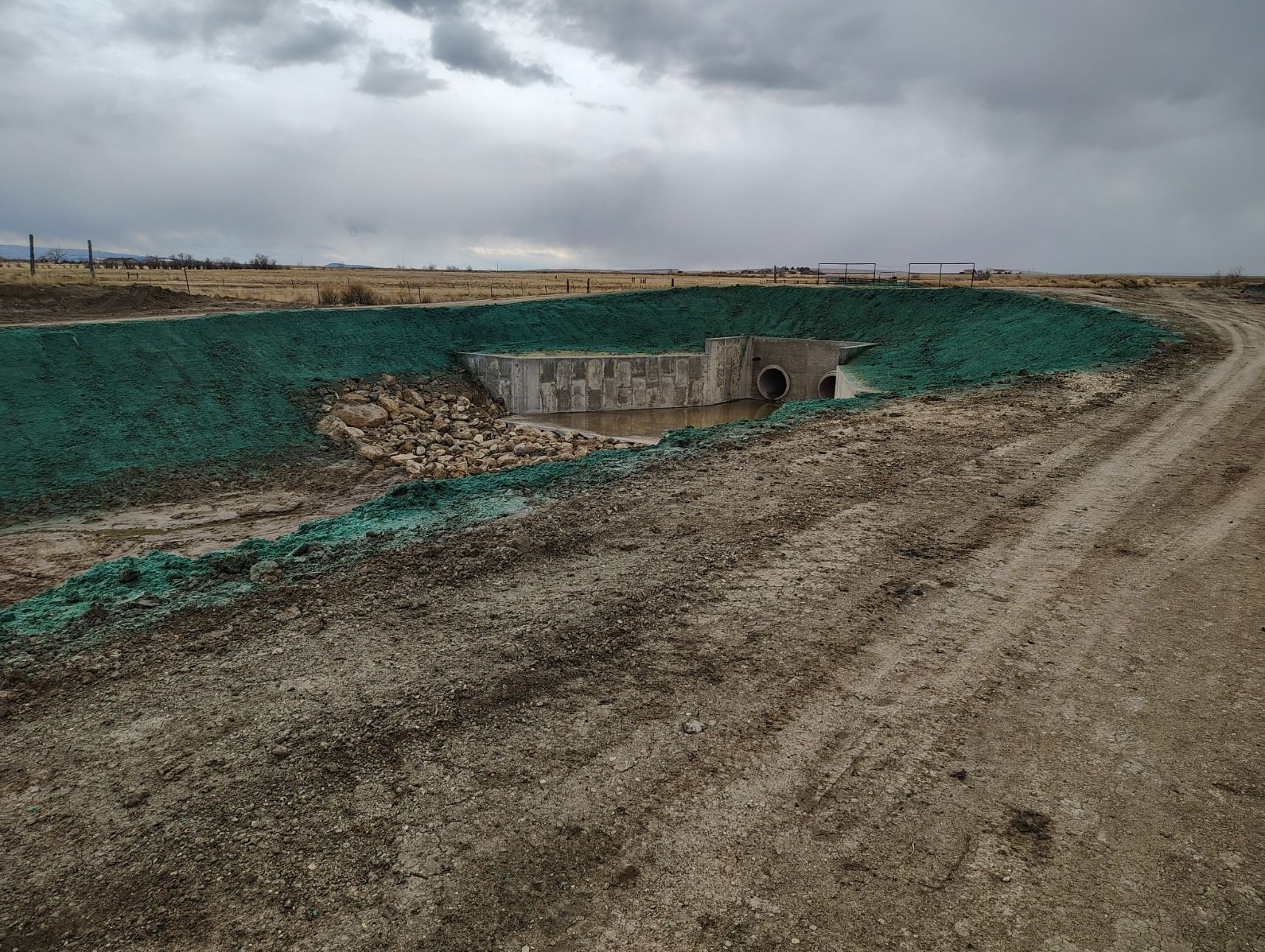 A concrete culvert headwall surrounded by a slope covered in green hydroseeding in a dry, rural field under a cloudy sky.