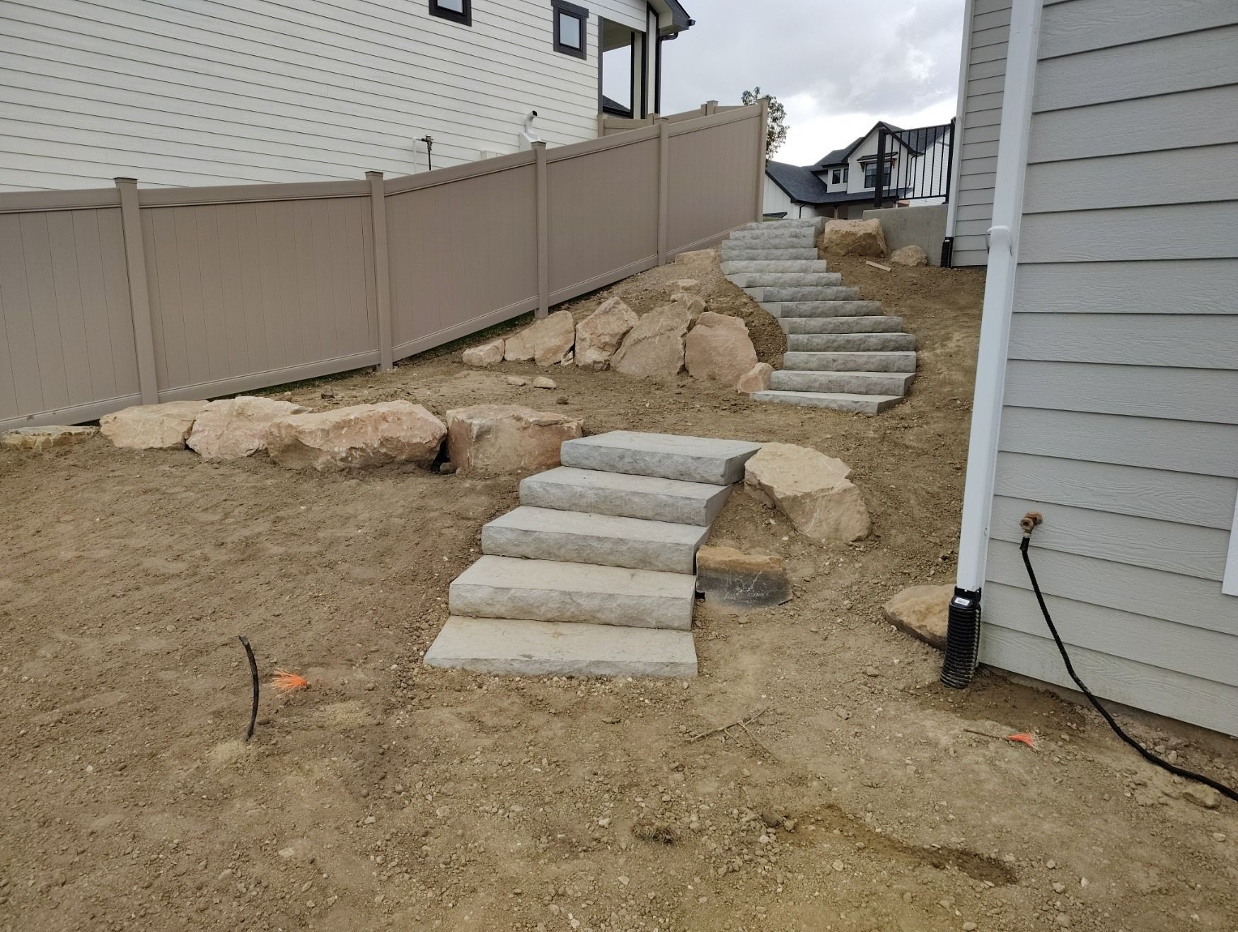 Two sets of stone steps leading up a sloped, dirt-covered backyard alongside a house and tan fence.