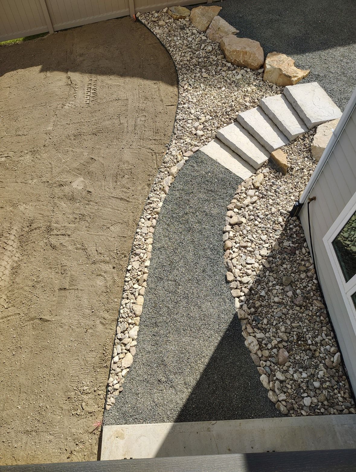 A curved path featuring grey stone, light gravel, and a set of concrete steps leading to the side of a building.