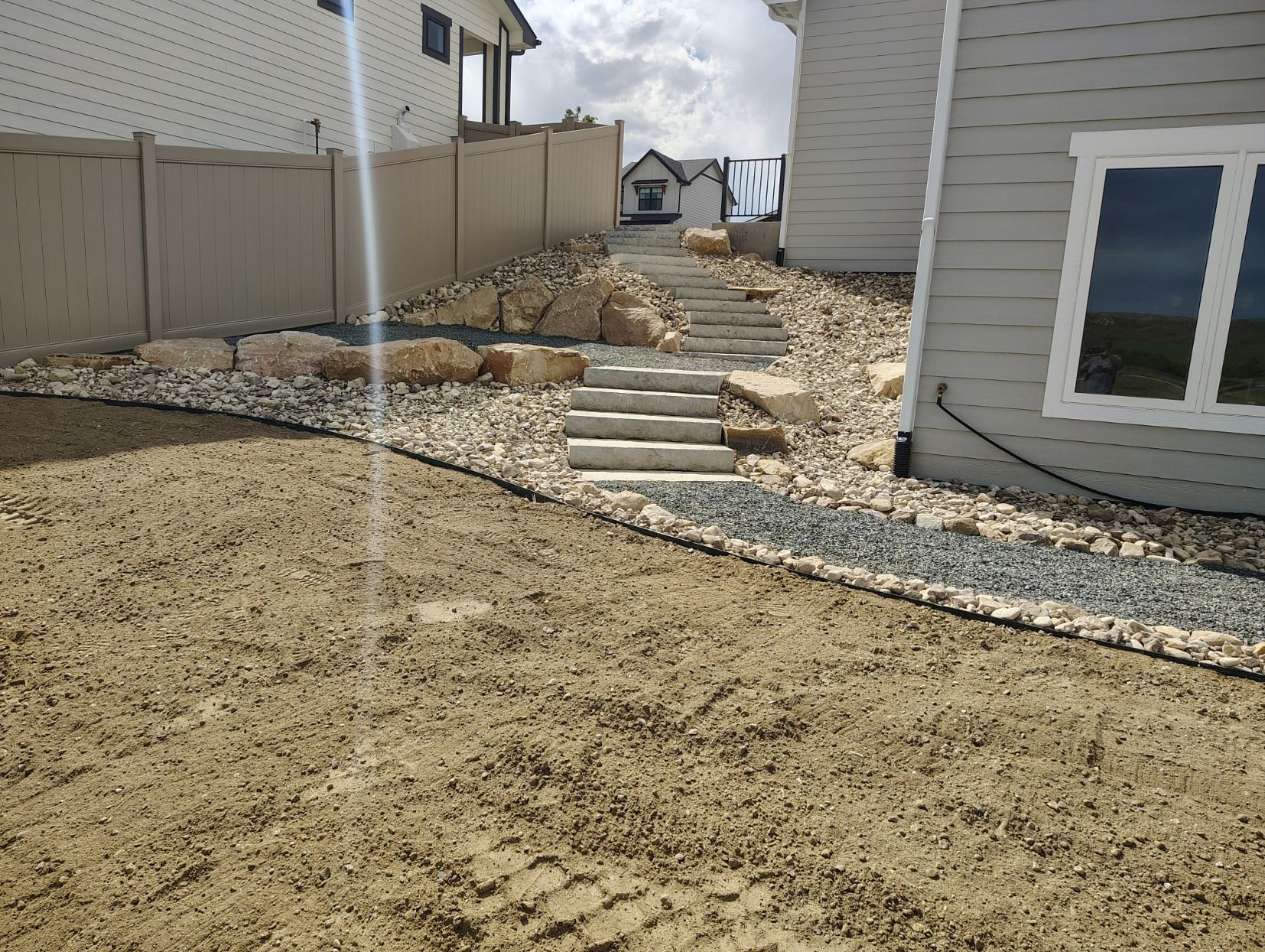 A backyard with a gravel path, stone stairs leading up a slope, tan vinyl fencing, and a gray house exterior.