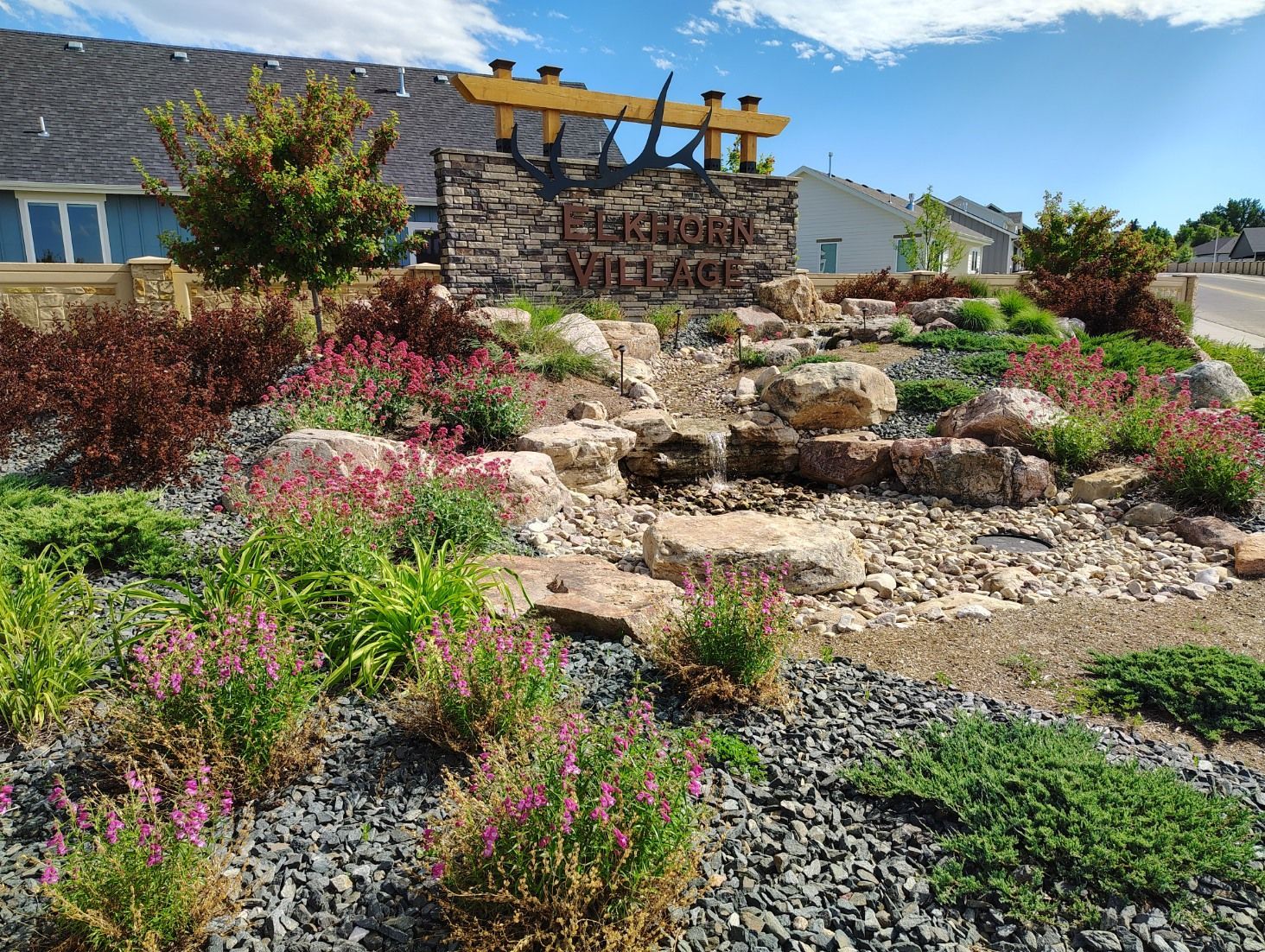 A stone sign for Heron Village sits in a landscaped garden with rocks, shrubs, and pink flowers against a blue sky.