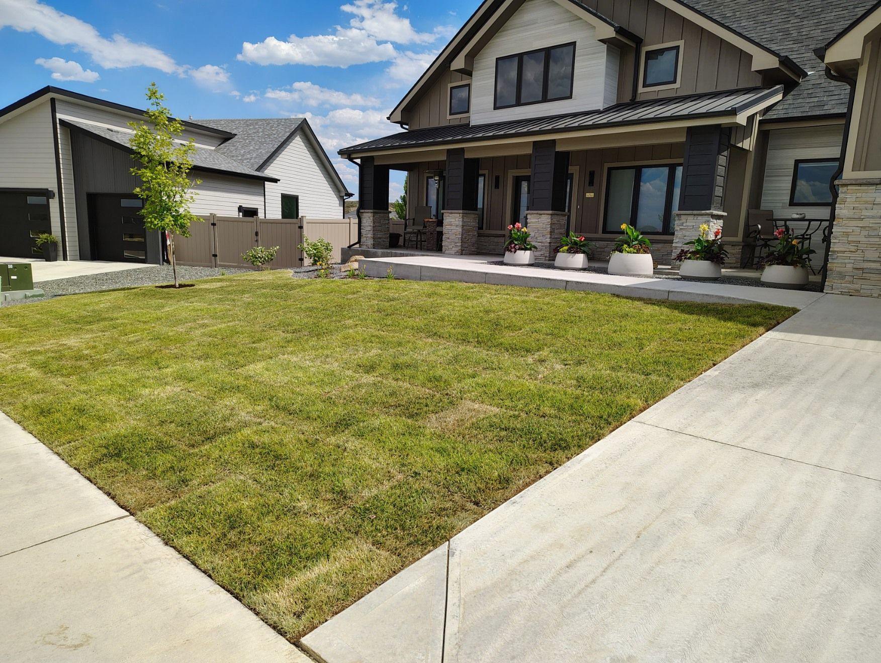 A front yard with a rectangular patch of grass bordered by concrete walkways leading to a modern house with stone columns.