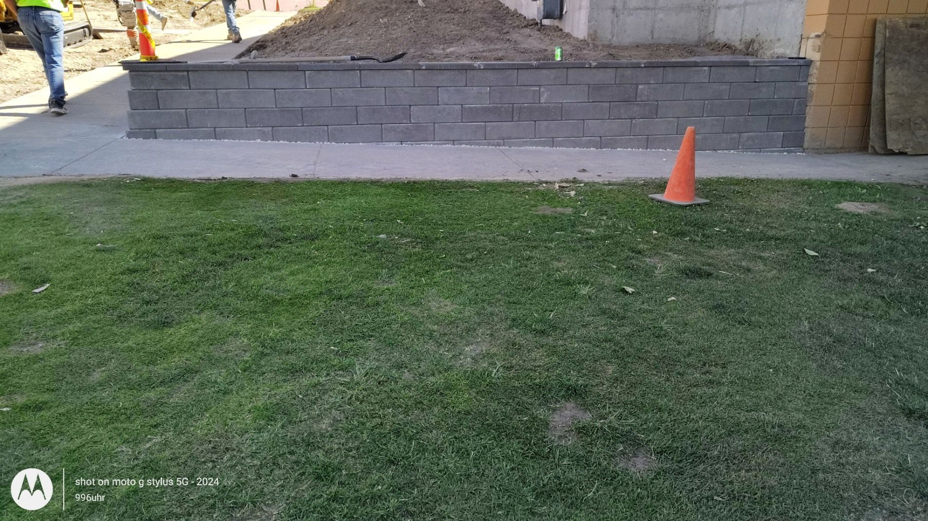 A newly built gray brick retaining wall sits next to a green lawn with a small orange safety cone placed on the grass.