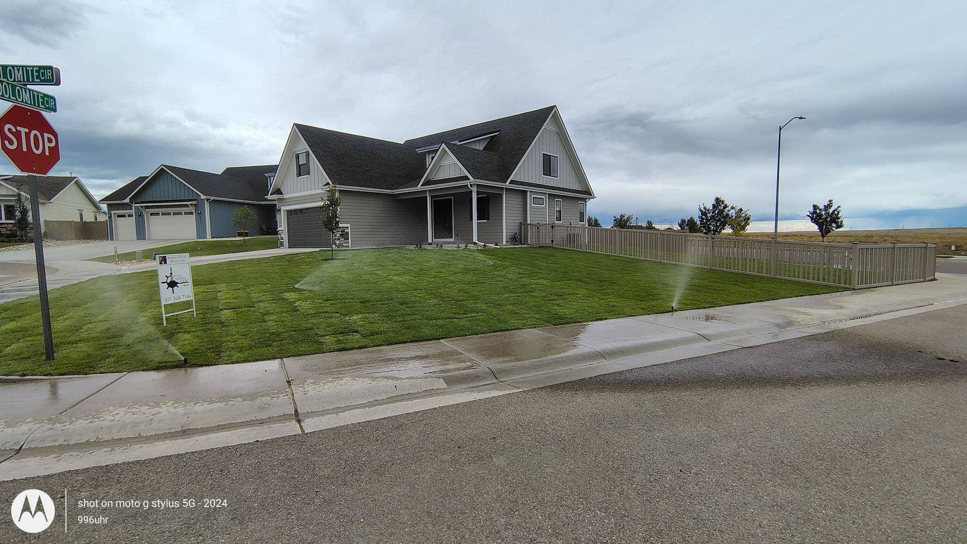 A suburban home on a corner lot with a sprinkler system watering the green grass under a cloudy sky.