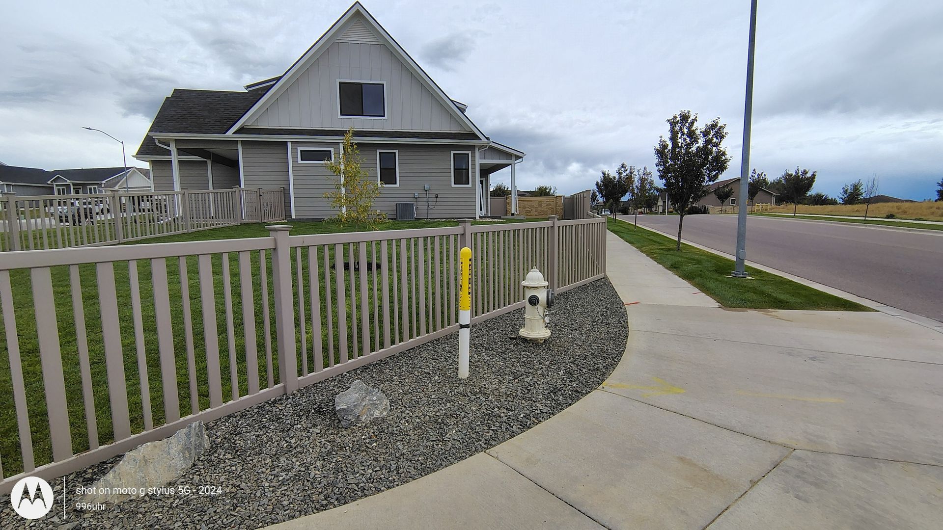 A light-colored house with a fence, a utility pole, and a fire hydrant beside a sidewalk on a cloudy day.