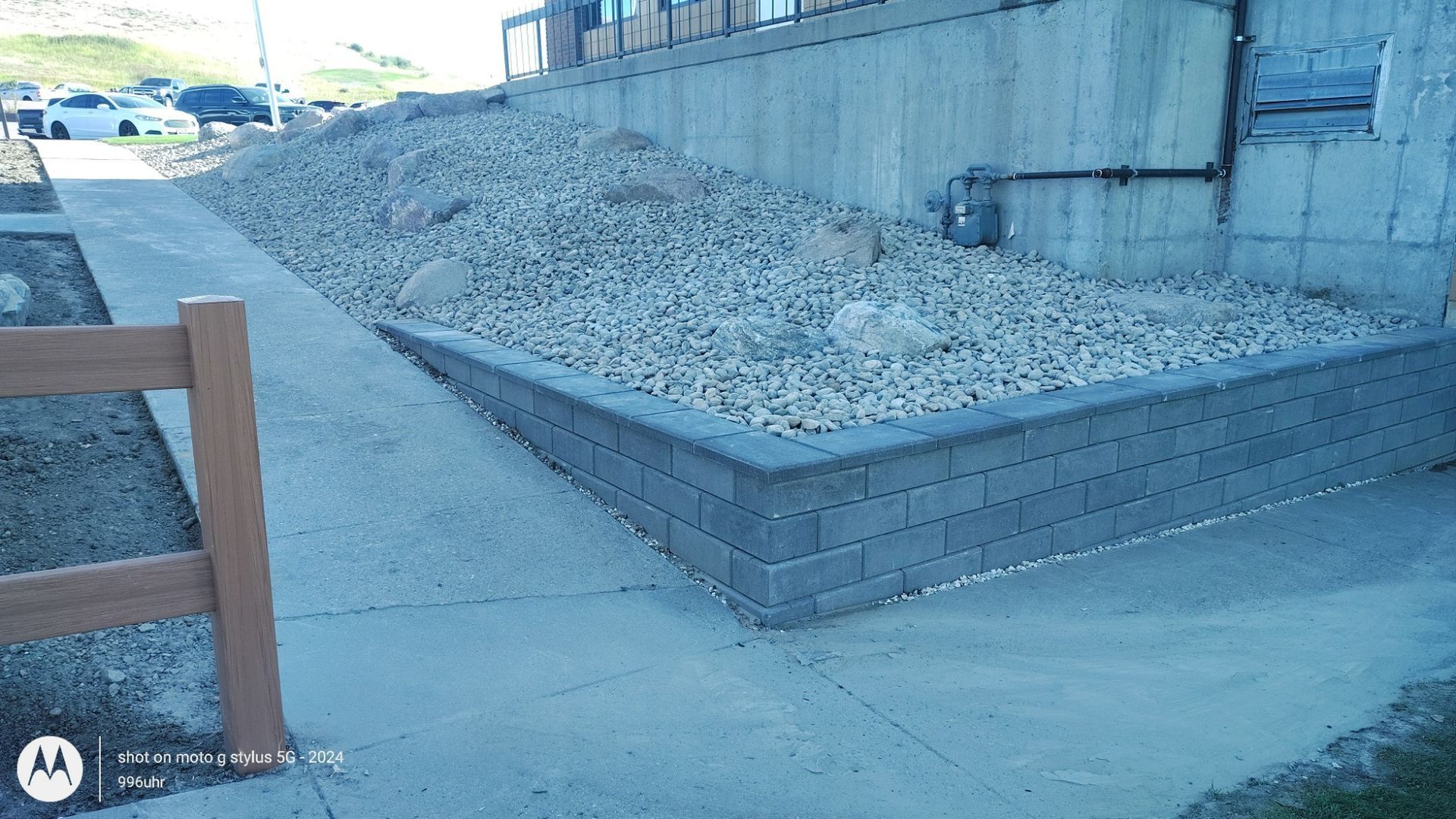 A concrete retaining wall with light-colored rocks, positioned next to a paved walkway and a wooden fence.