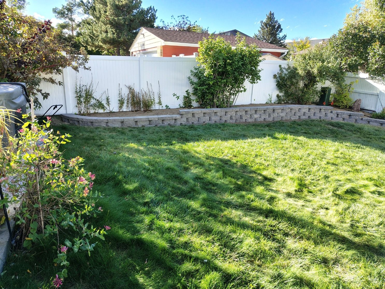 A backyard featuring a stone retaining wall in front of a white privacy fence, surrounded by green grass and bushes.