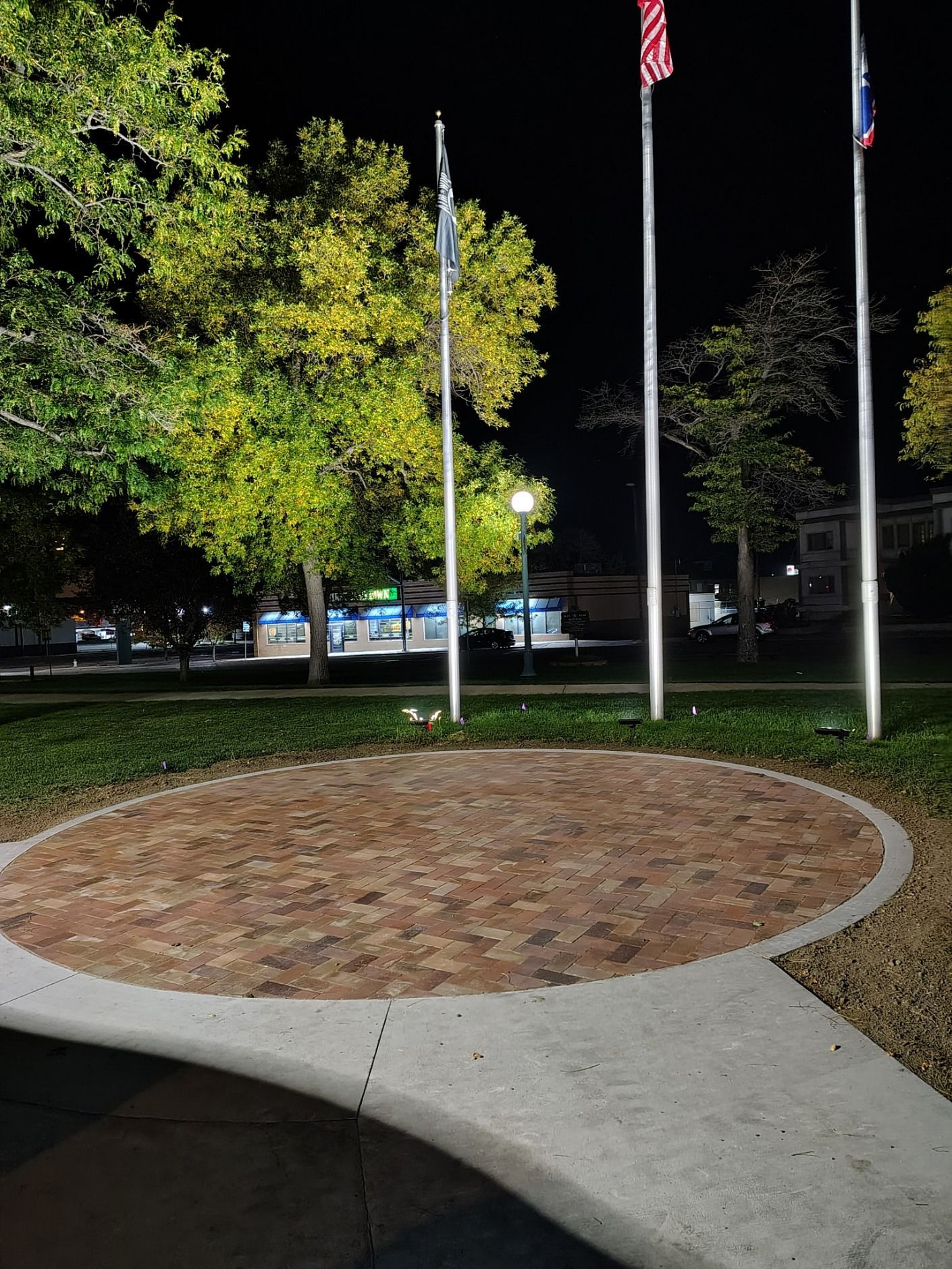 Three flagpoles rise behind a circular brick plaza illuminated by nearby park lights at night.