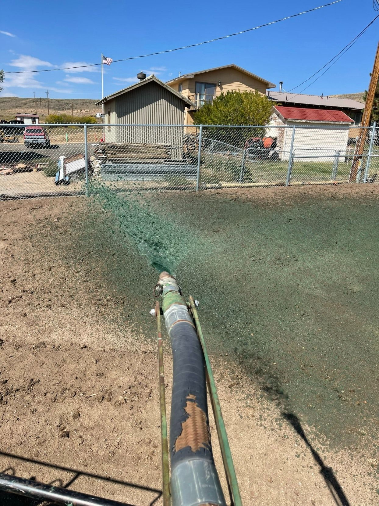 A hose sprays green hydroseed mixture over a bare patch of dirt in a residential yard near a chain-link fence.