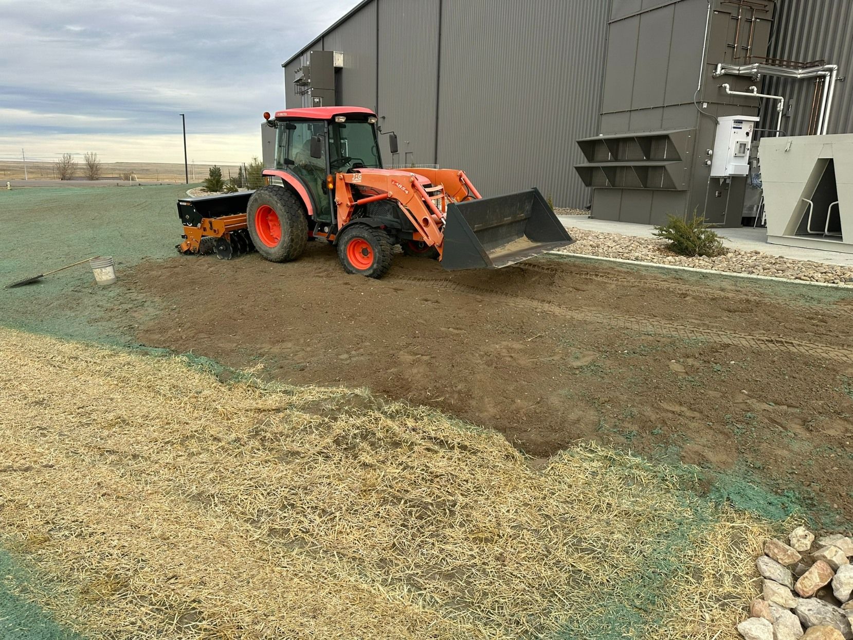 An orange tractor with a front loader moves soil near a large industrial building on a gravel-covered site.