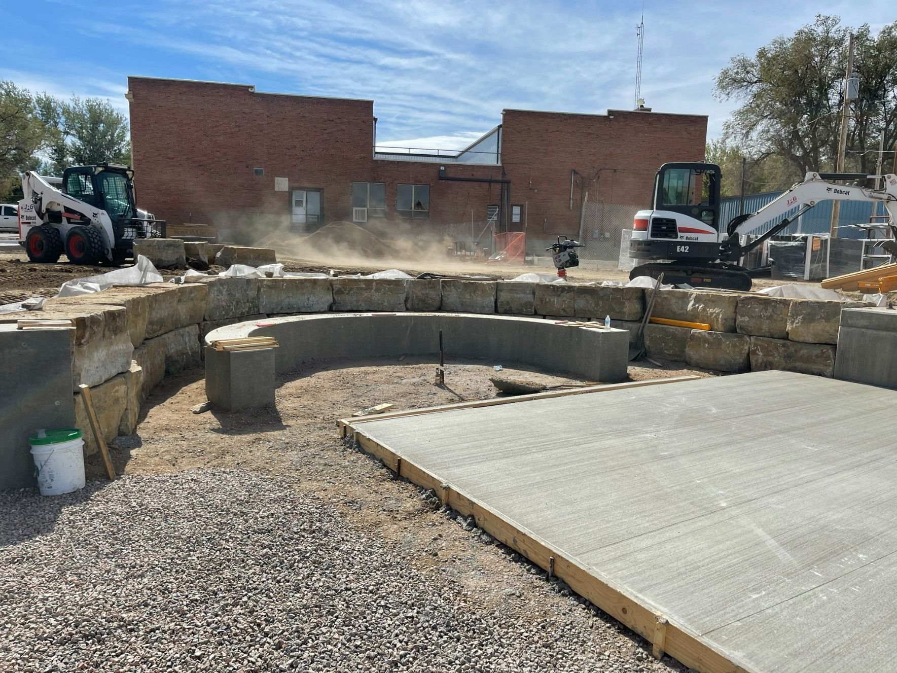 Construction site with a stone retaining wall, concrete pad, and heavy machinery in front of a brick building.