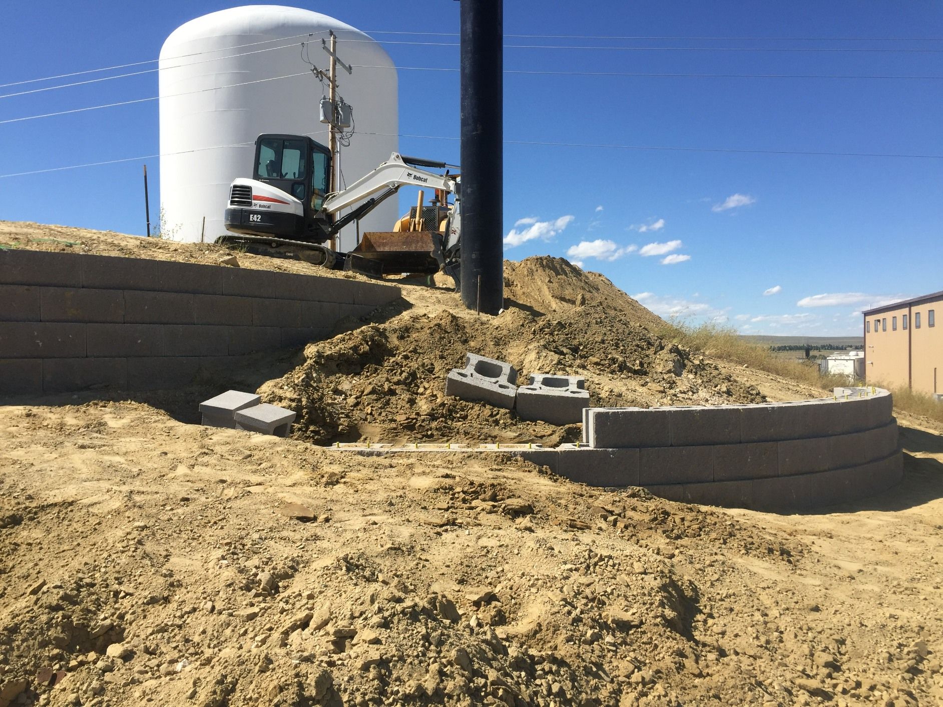A construction excavator works on a dirt hill beside a white storage tank and a new concrete retaining wall.