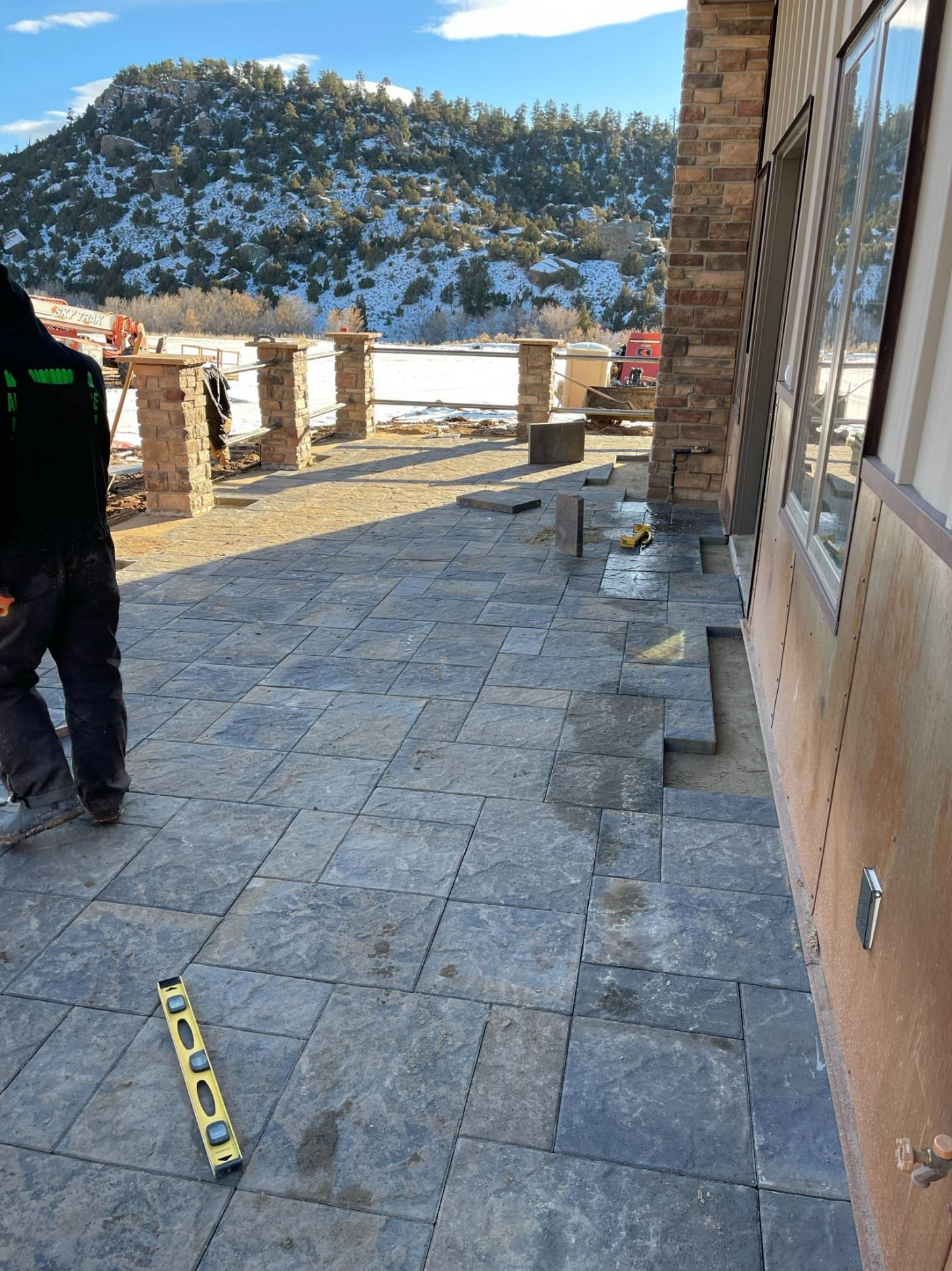 A worker stands on a partially installed stone patio outside a building, with snowy mountains in the background.