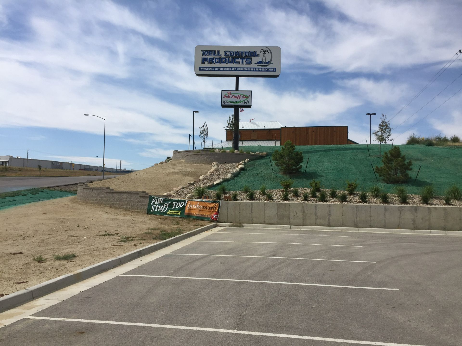 A large sign for a business stands above a landscaped hill next to a parking lot under a blue sky with scattered clouds.